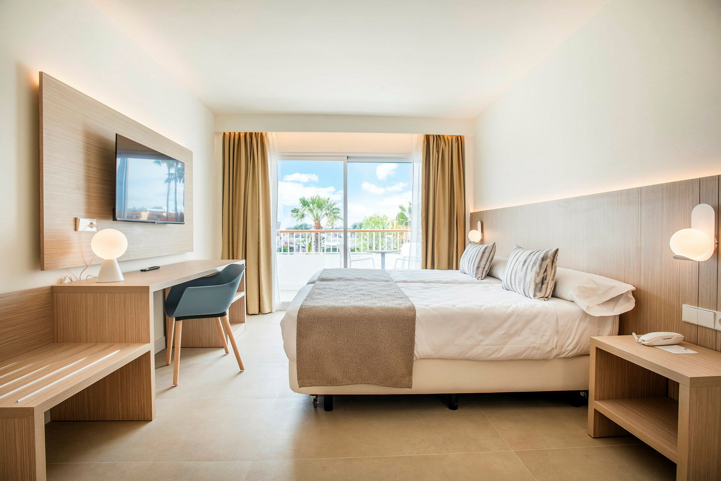 Modernt inrett hotellrum med säng, skrivbord och stora glasdörrar mot en balkong. (AI-genererad text)