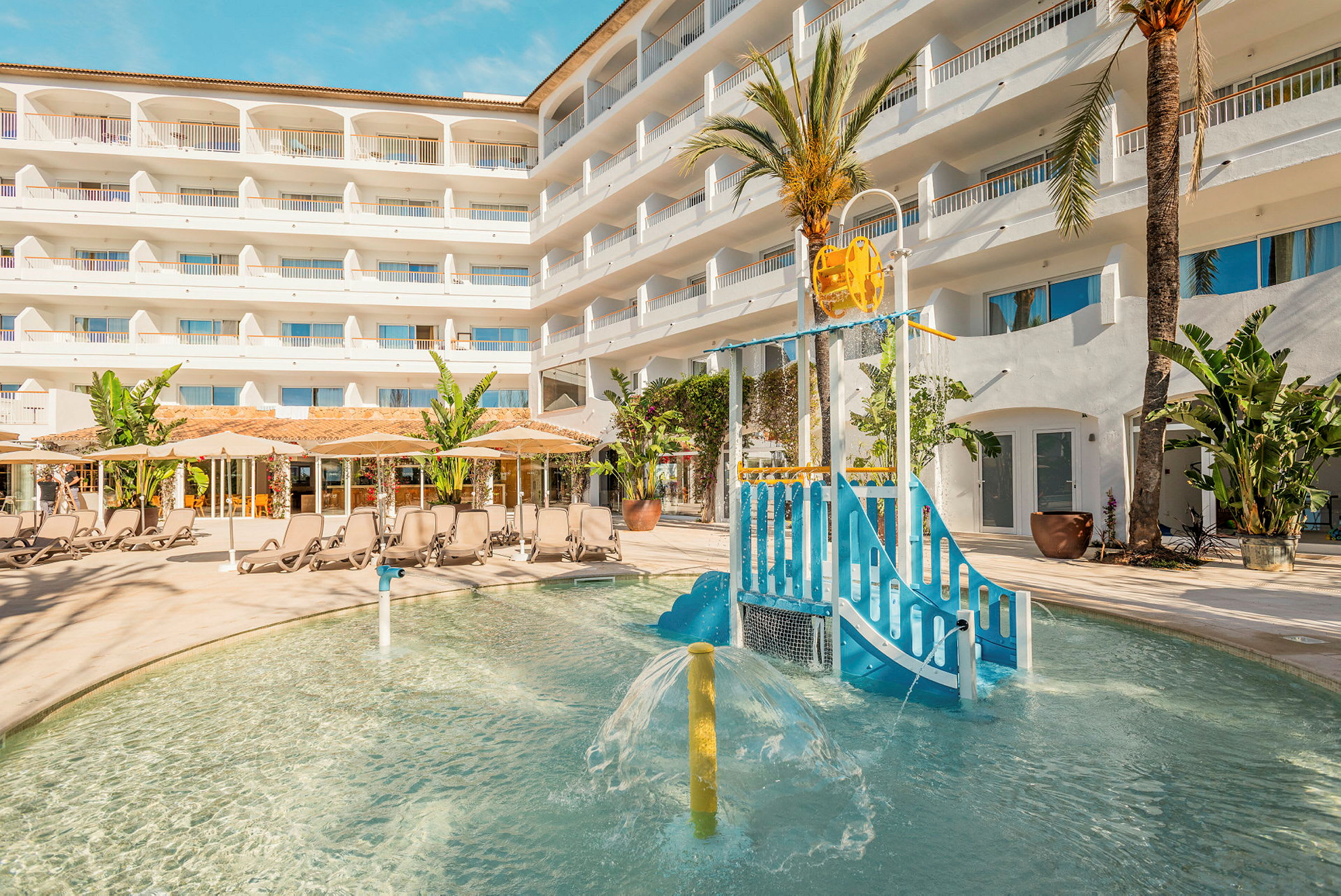 Utomhuspoolområde med barnlekplats och solstolar framför hotellbyggnad. (AI-genererad text)