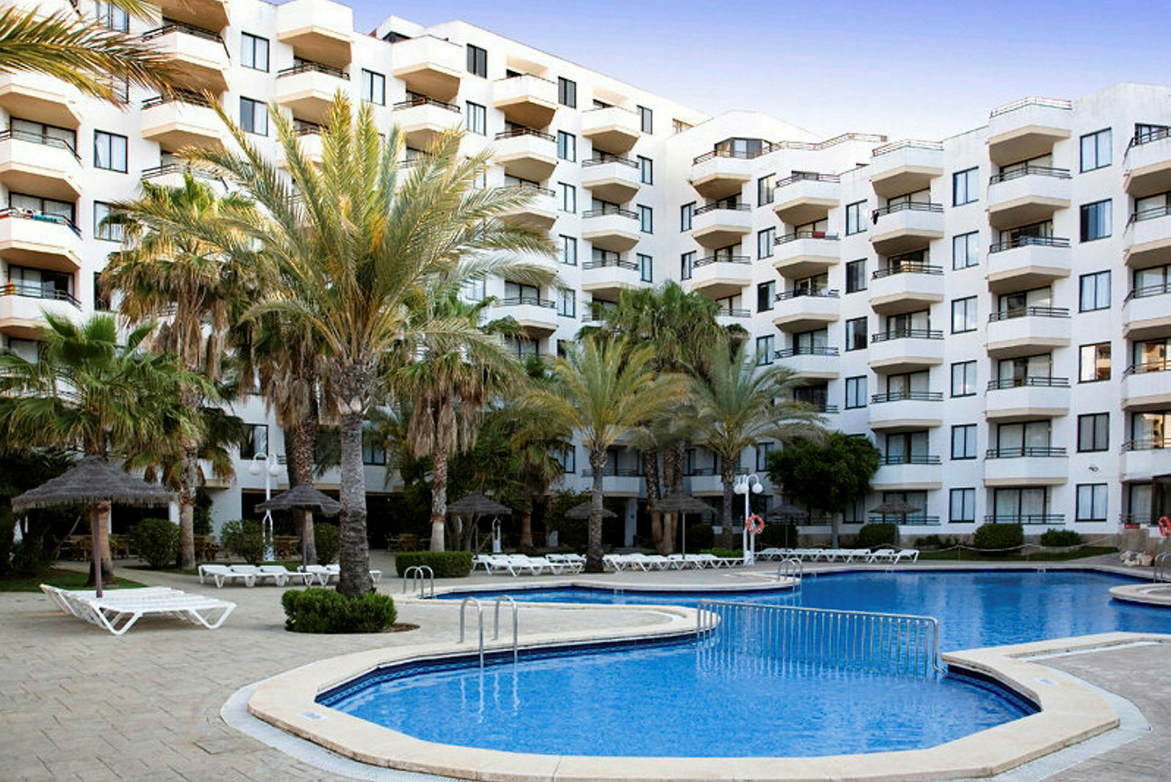 Hotellbyggnad med flera balkonger, omgiven av palmträd och en poolområde med solstolar. (AI-genererad text)