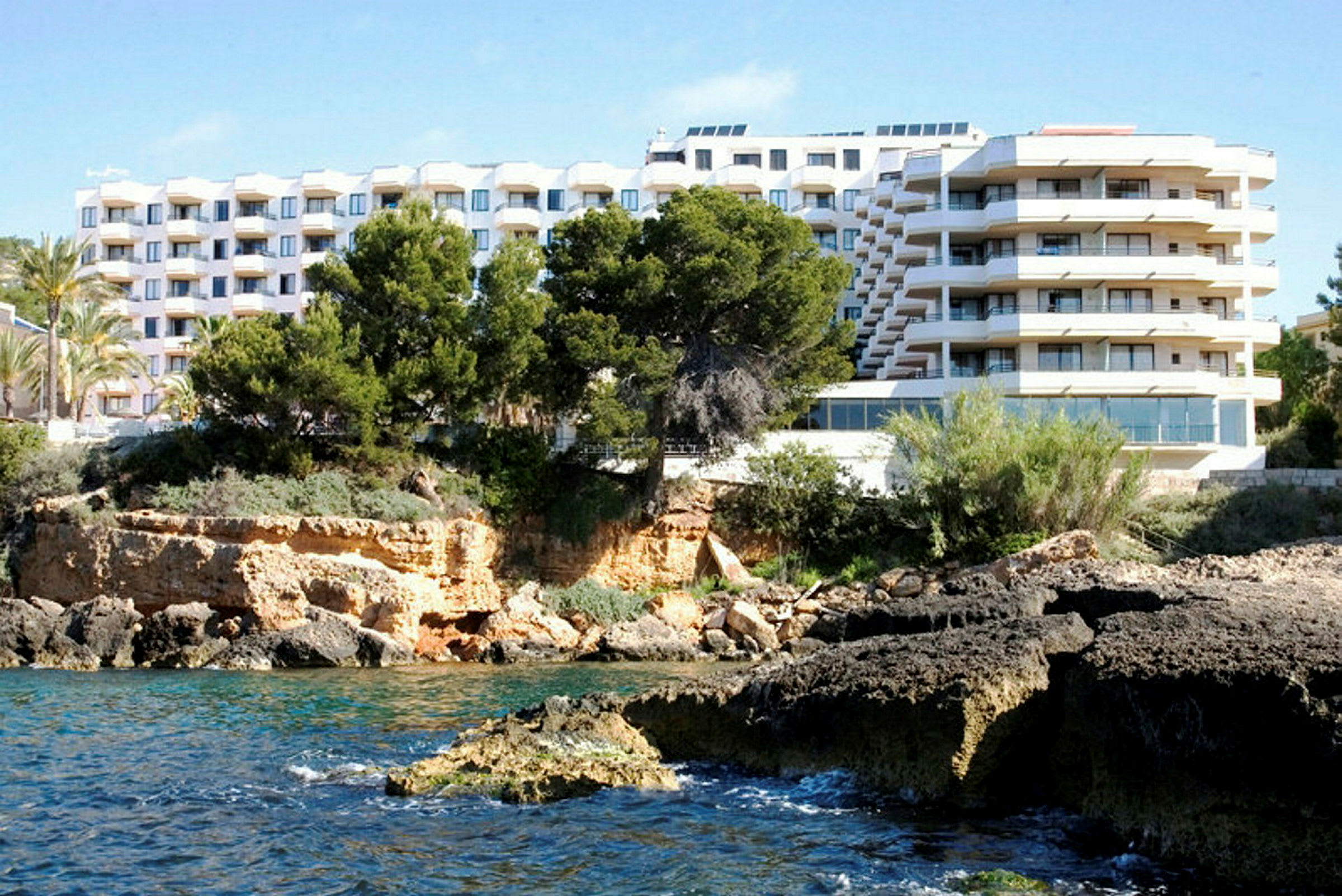 Ett hotellkomplex med flera våningar är beläget intill en stenig kust med träd och buskar. (AI-genererad text)