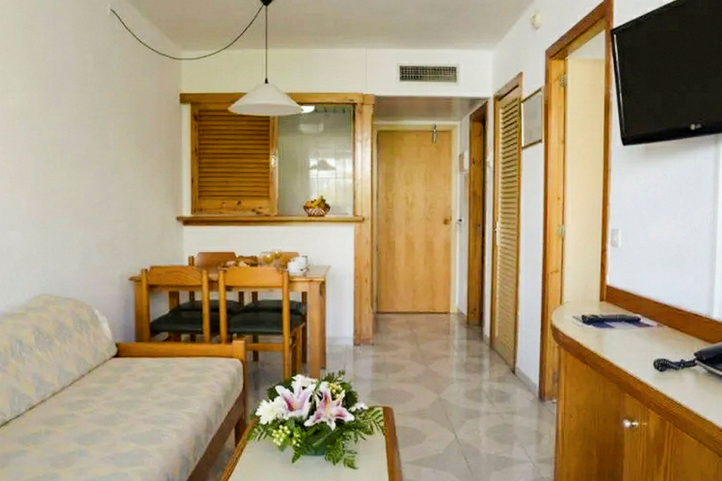 Rummet består av en sittgrupp med en soffa och ett soffbord, en matplats med bord och stolar samt en tv på väggen. Det finns dörrar som leder till andra delar av boendet. (AI-genererad text)