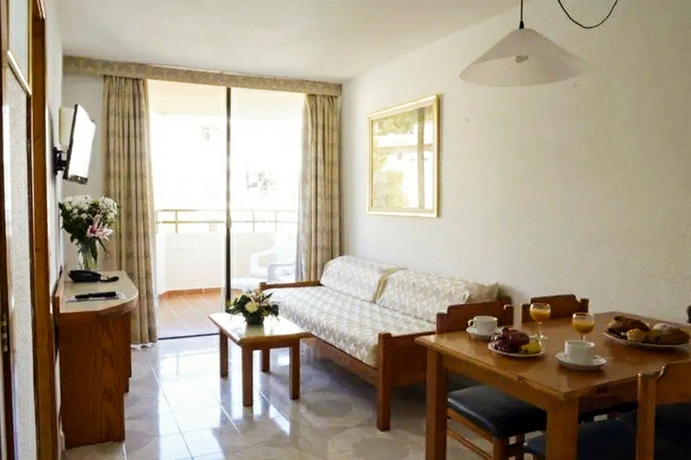 En lägenhet med ett vardagsrum som innehåller en soffa, ett bord med blommor, en matplats med dukat bord och en balkong i bakgrunden. (AI-genererad text)