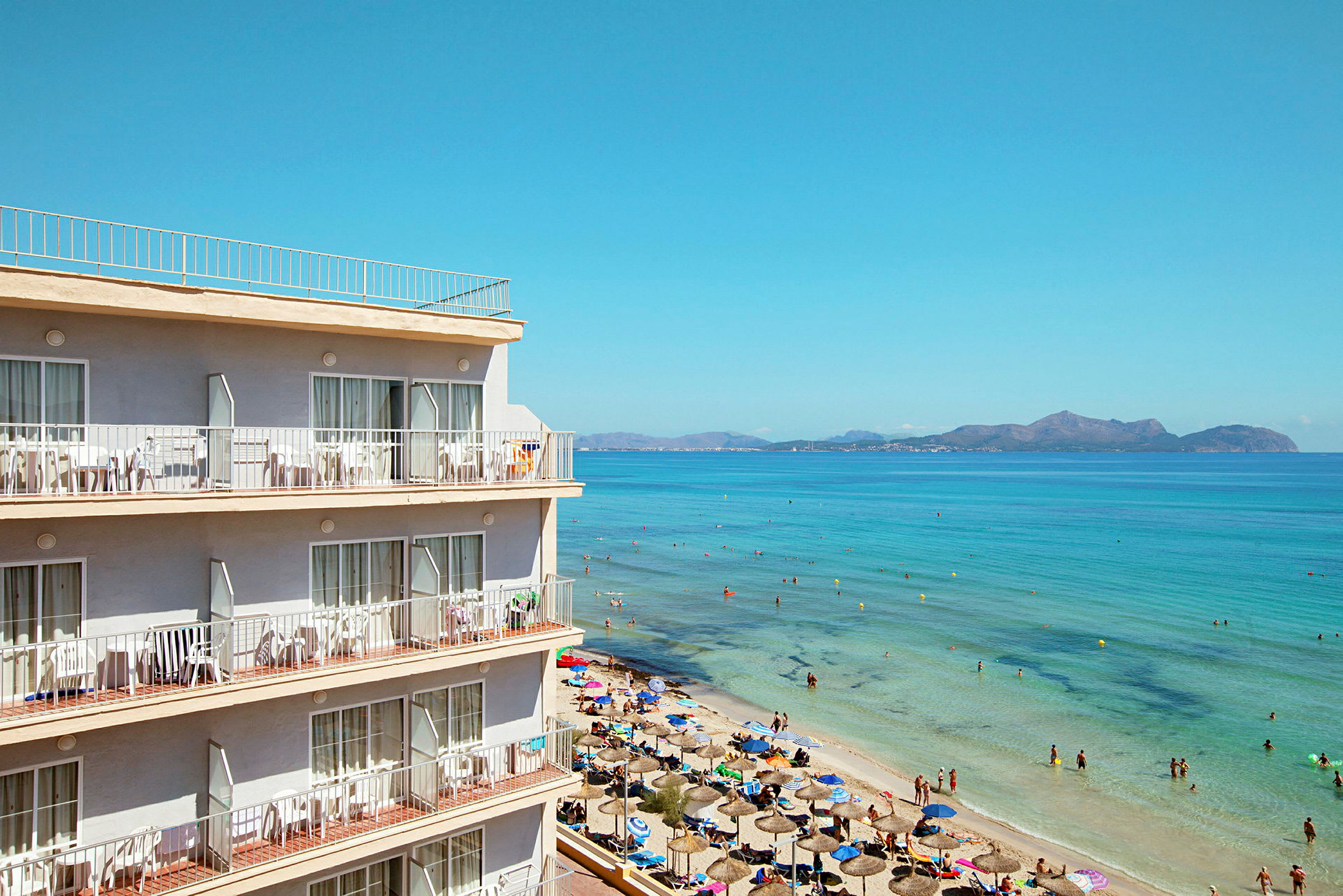 Ett strandhotell nära en sandstrand med paraplyer, och personer badar i klart blått vatten. (AI-genererad text)