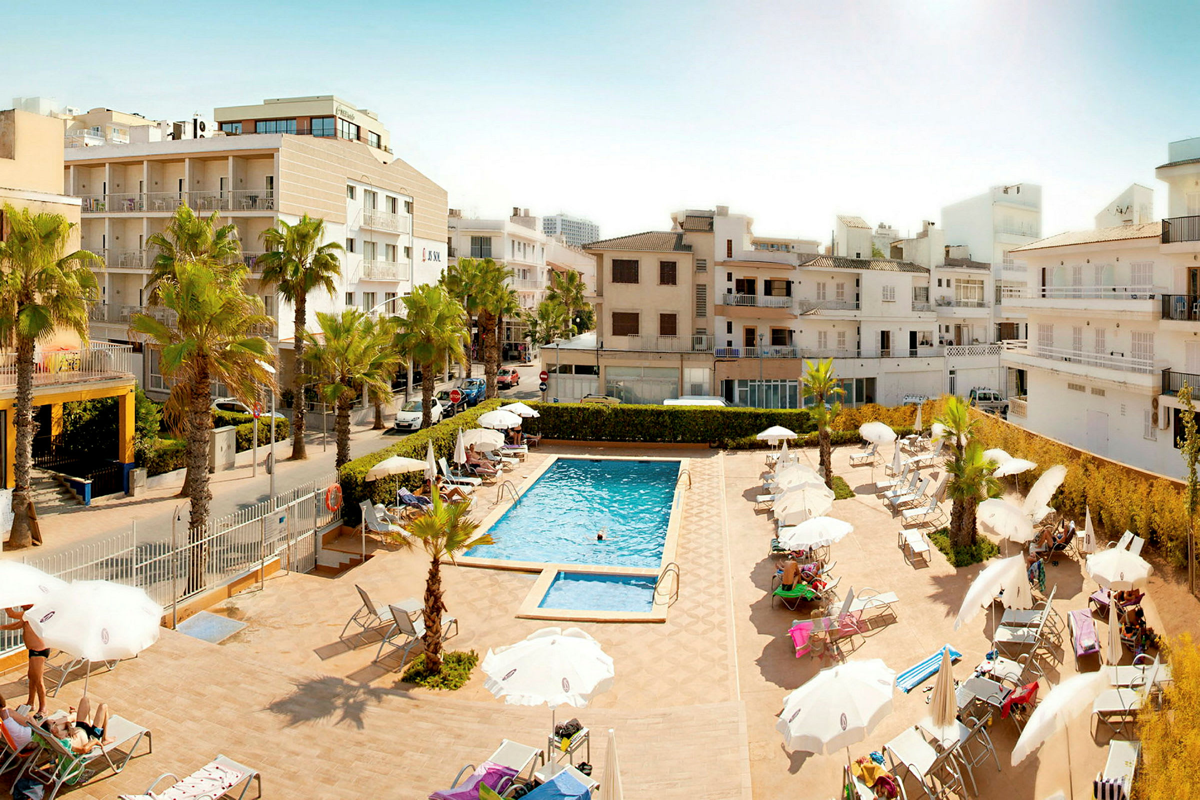 Hotellets poolområde med solstolar och parasoller omgivna av byggnader. (AI-genererad text)