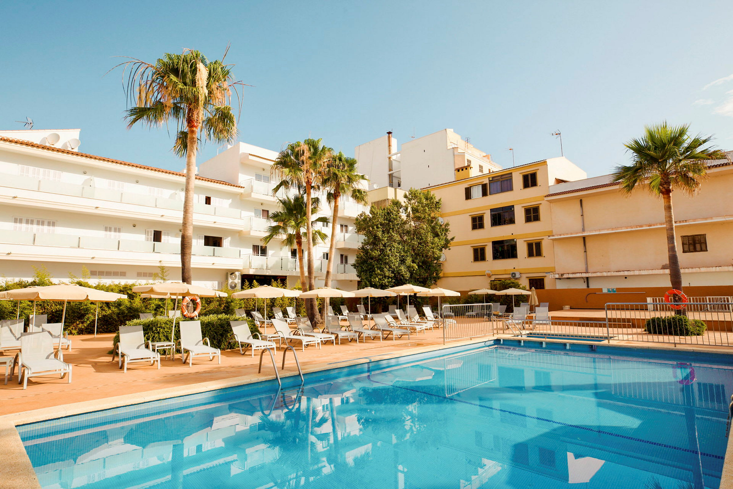 Exteriör av hotell med pool och solstolar, omgiven av byggnader och palmer. (AI-genererad text)