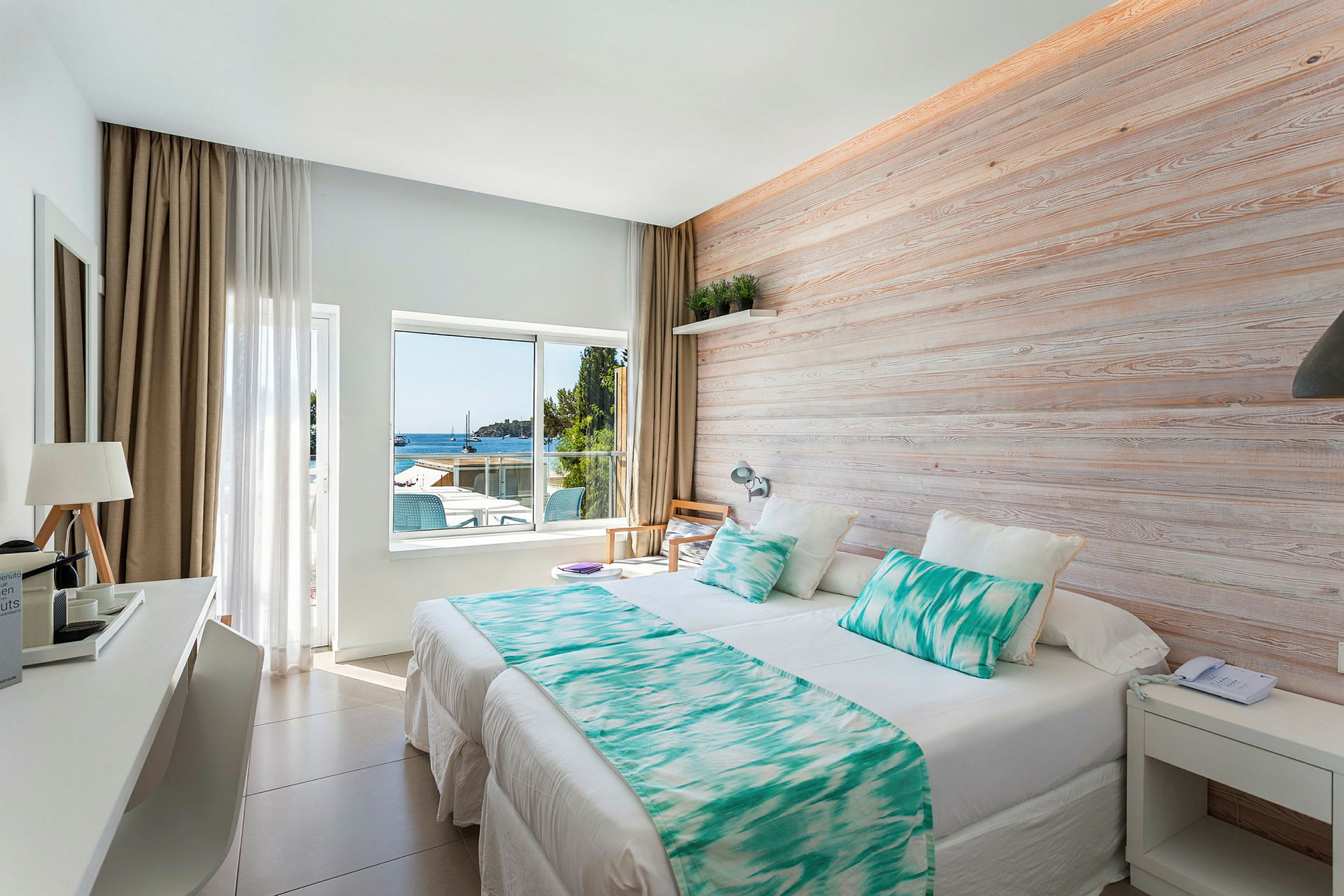 Ett ljust hotellrum med en stor säng, träpanel på väggen och ett fönster som vetter mot havet. (AI-genererad text)