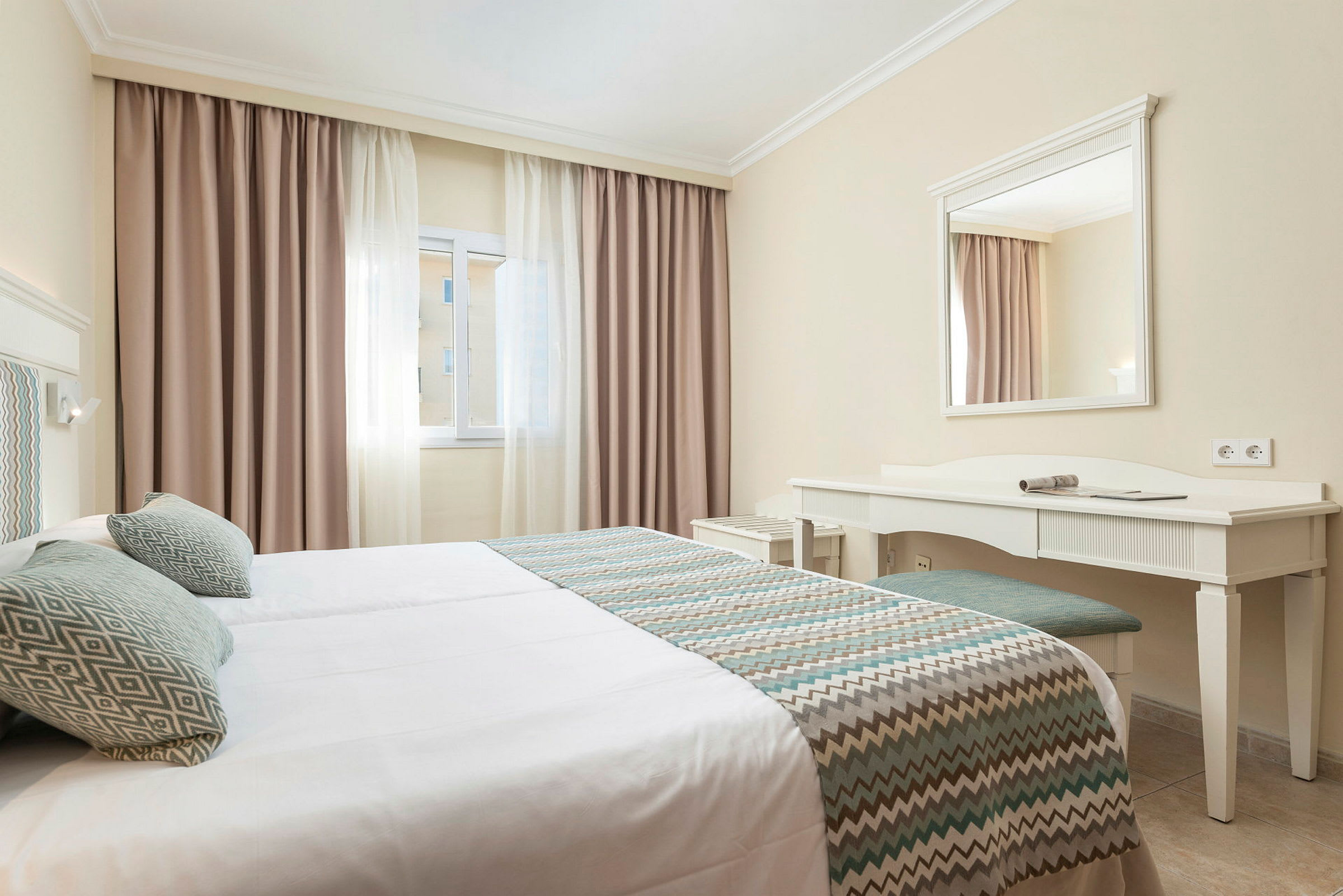 Hotelrum med enkelsängar, fönster med ljusa gardiner samt ett skrivbord med spegel. (AI-genererad text)