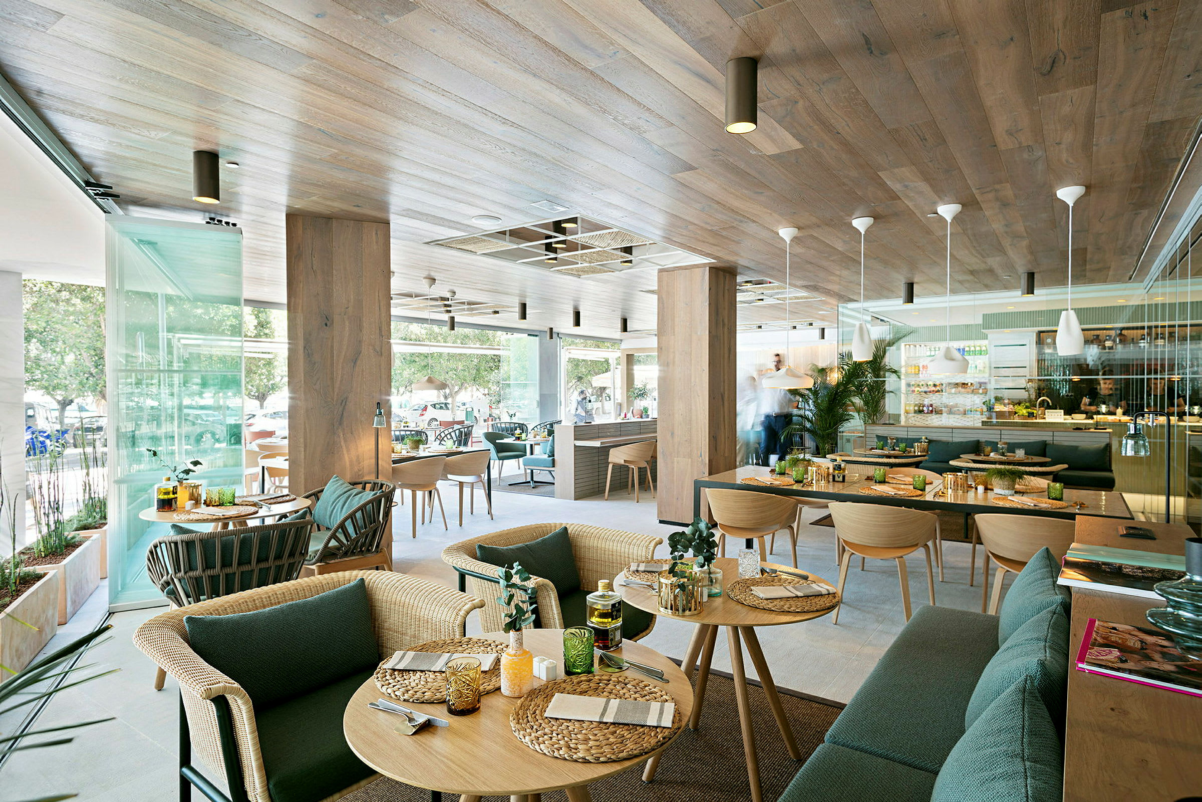 En modern restaurang med träinredning och stora glasfönster. Det finns bord med serverad mat och inredning med gröna växter. (AI-genererad text)