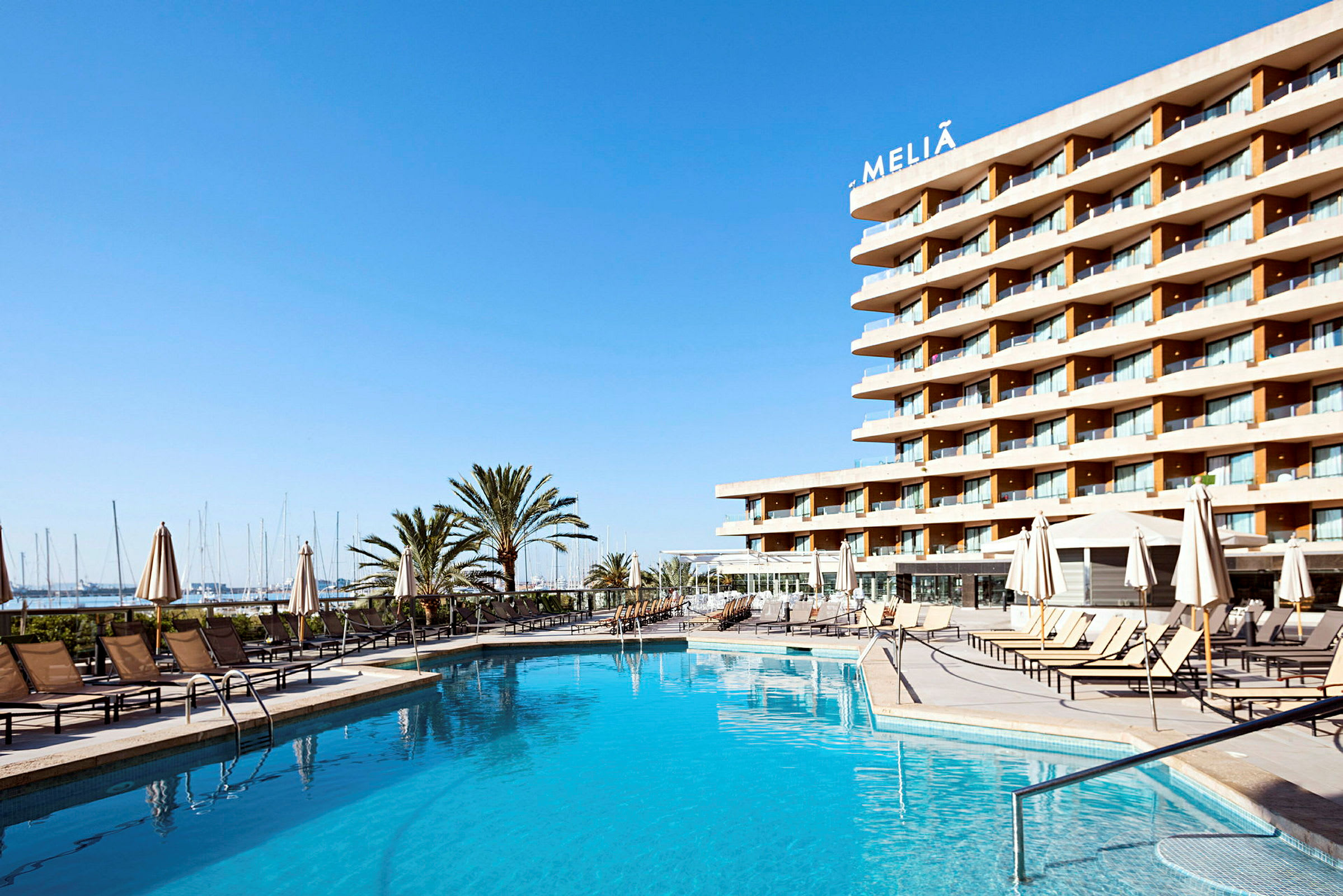 Ett hotell med en pool i förgrunden, omgiven av solstolar och parasoller, med havet och en småbåtshamn i bakgrunden under en klarblå himmel. (AI-genererad text)