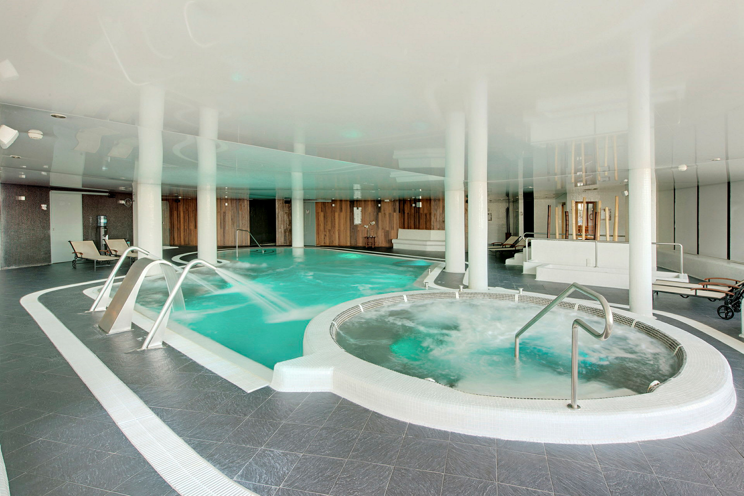 En inomhuspool med en jacuzzi bredvid, omgiven av vita pelare och liggstolar. (AI-genererad text)