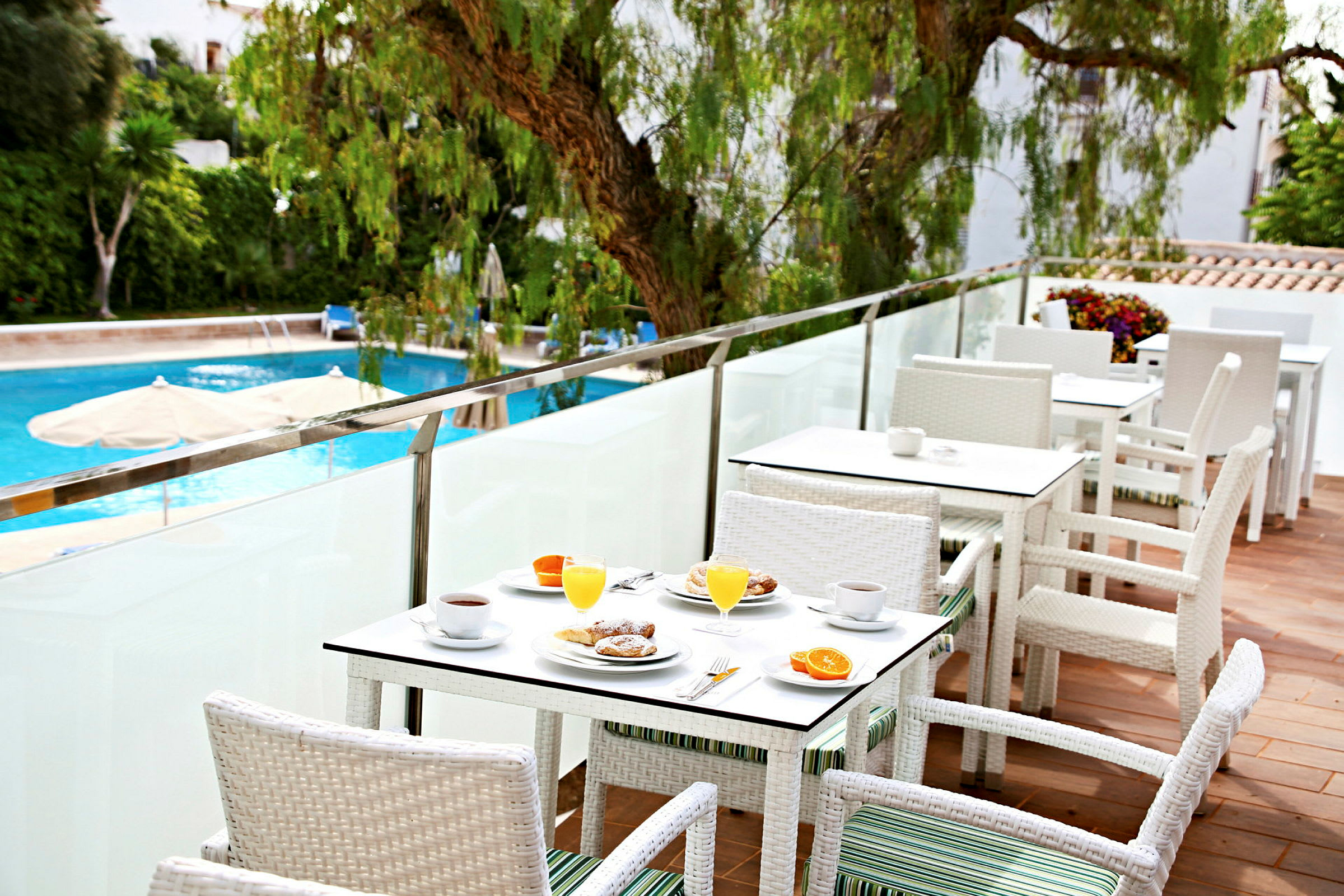 Uteservering med vita bord och stolar, frukost serverad på borden, och en swimmingpool i bakgrunden. (AI-genererad text)
