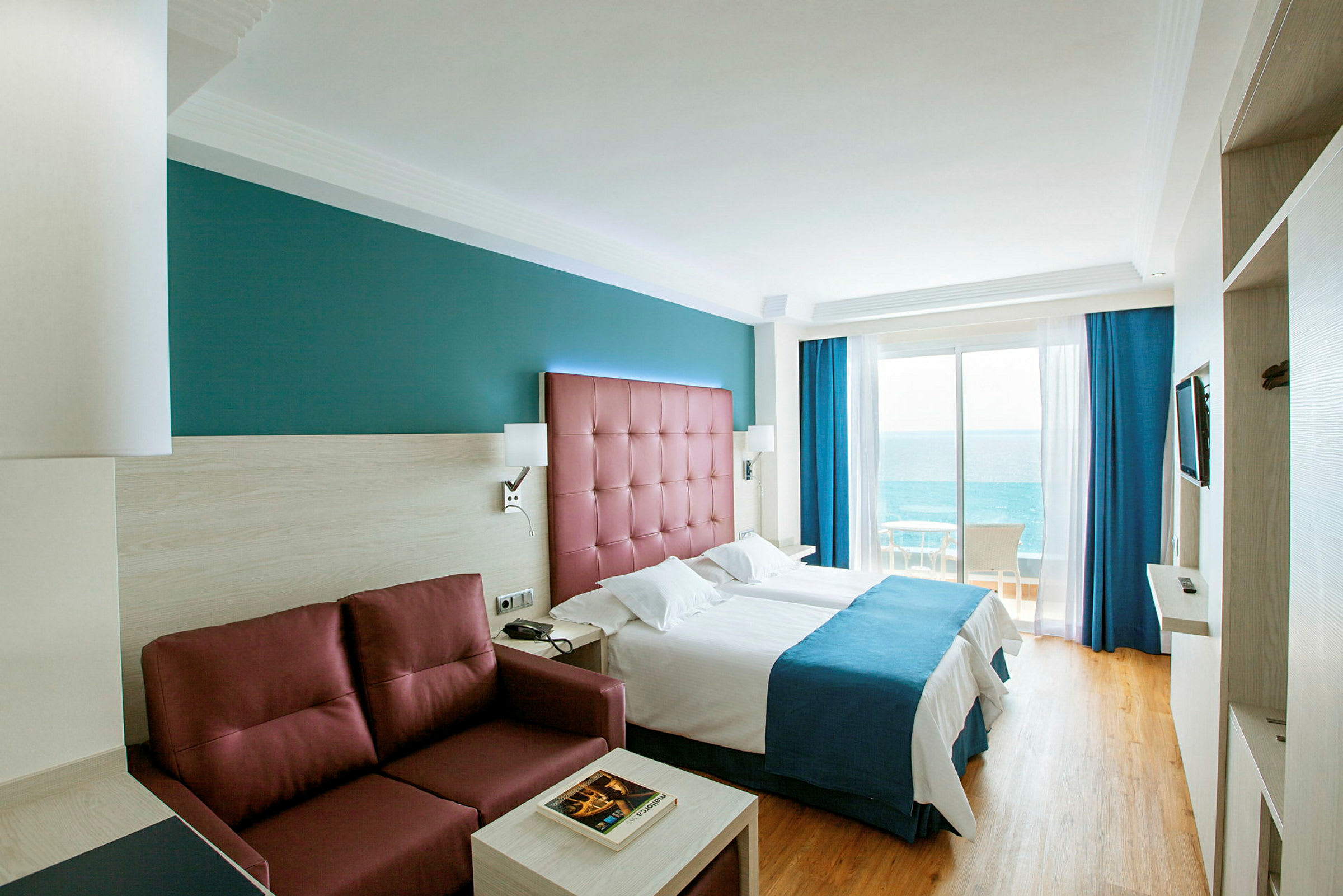 Hotellrum med två enkelsängar, en röd soffa och havsutsikt från balkongen. (AI-genererad text)