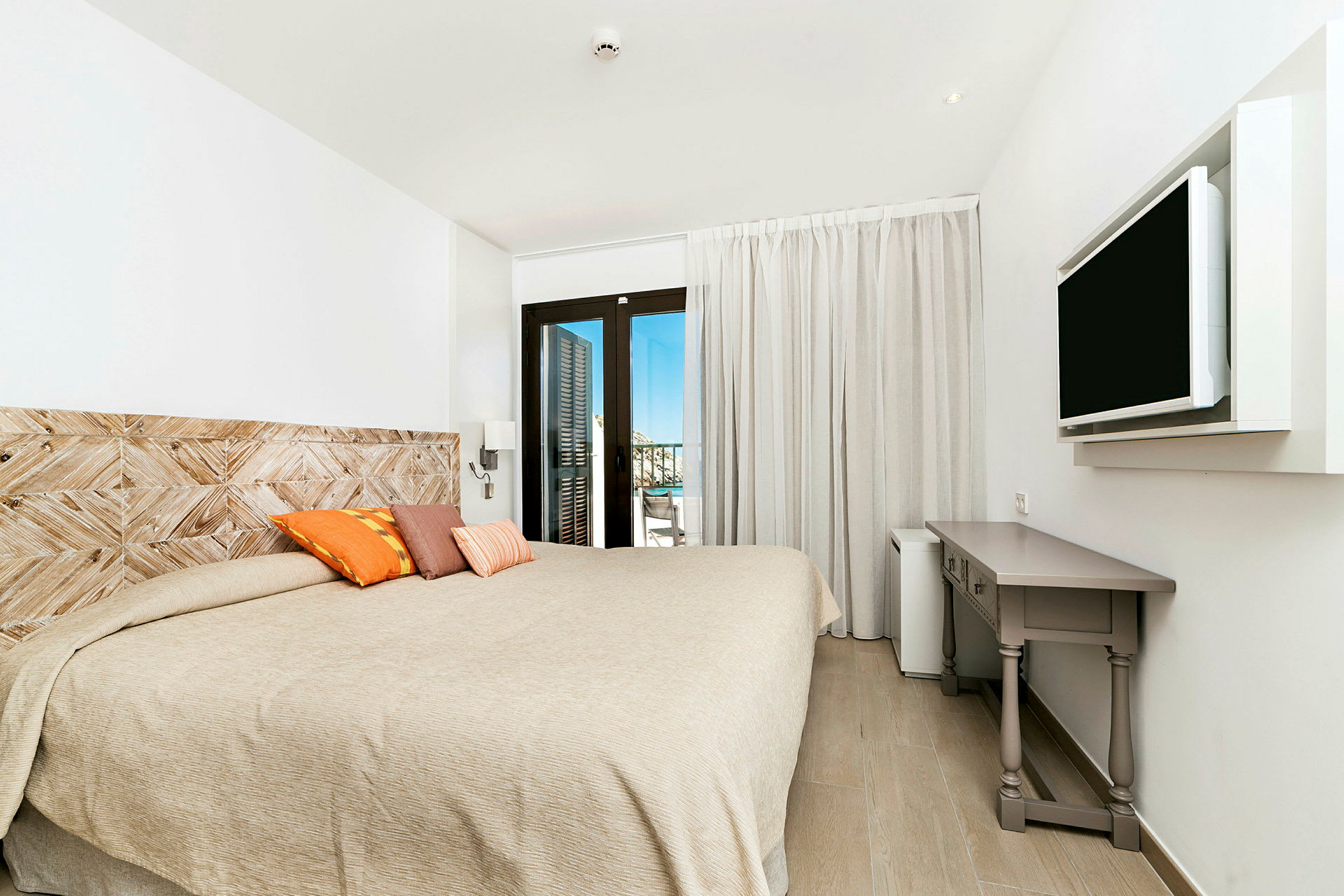 Ett hotellrum med en stor säng, dekorativa kuddar, en väggmonterad TV, och ett skrivbord. Balkongdörrar leder till en utsikt. (AI-genererad text)