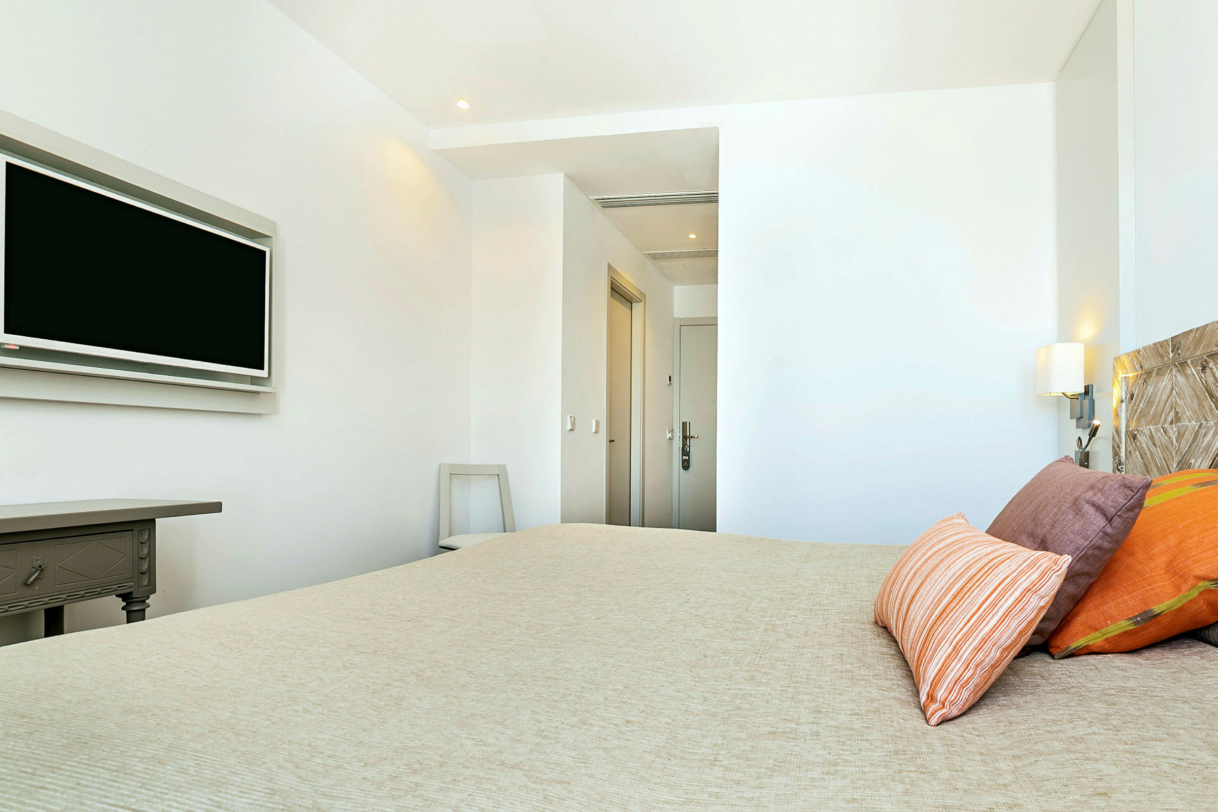 Ett hotellrum med en säng, dekorativa kuddar och en väggmonterad TV. Ett bord och dörrar syns också. (AI-genererad text)