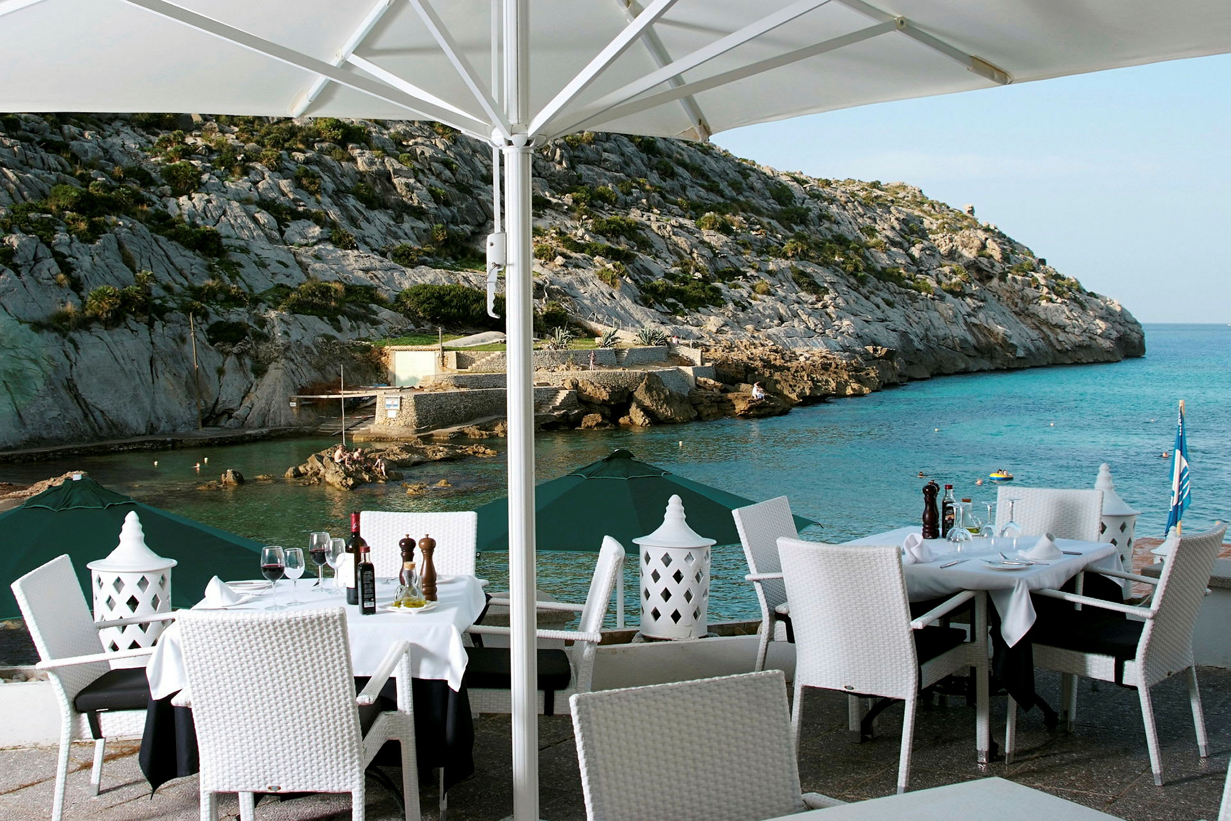 En utomhusrestaurang med dukade bord och vita stolar med utsikt över ett klippigt kustlandskap och havet. (AI-genererad text)