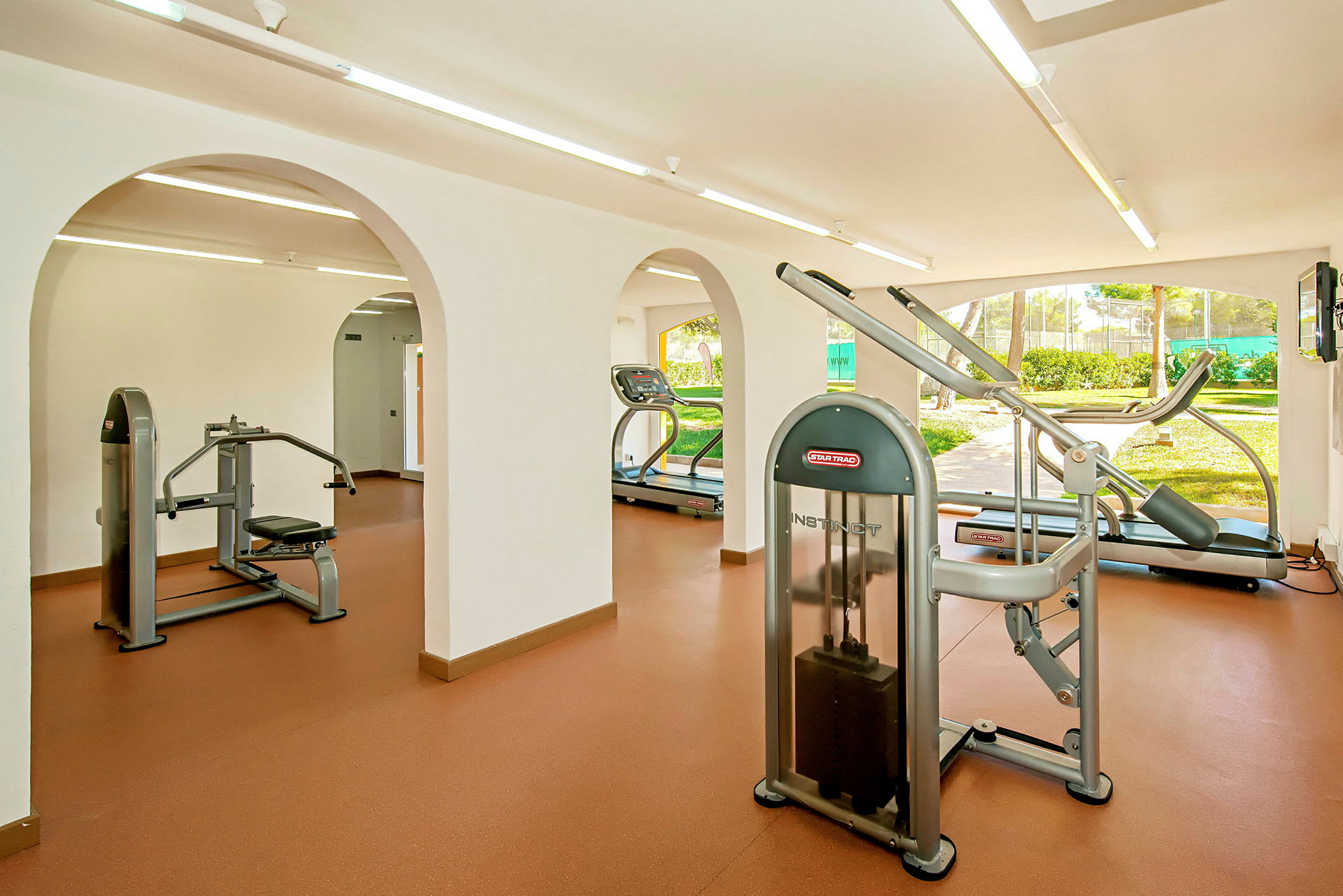 Ett gym med olika träningsmaskiner som löpband och styrketräningsutrustning. Rummet har stora fönster med utsikt mot grönska. (AI-genererad text)