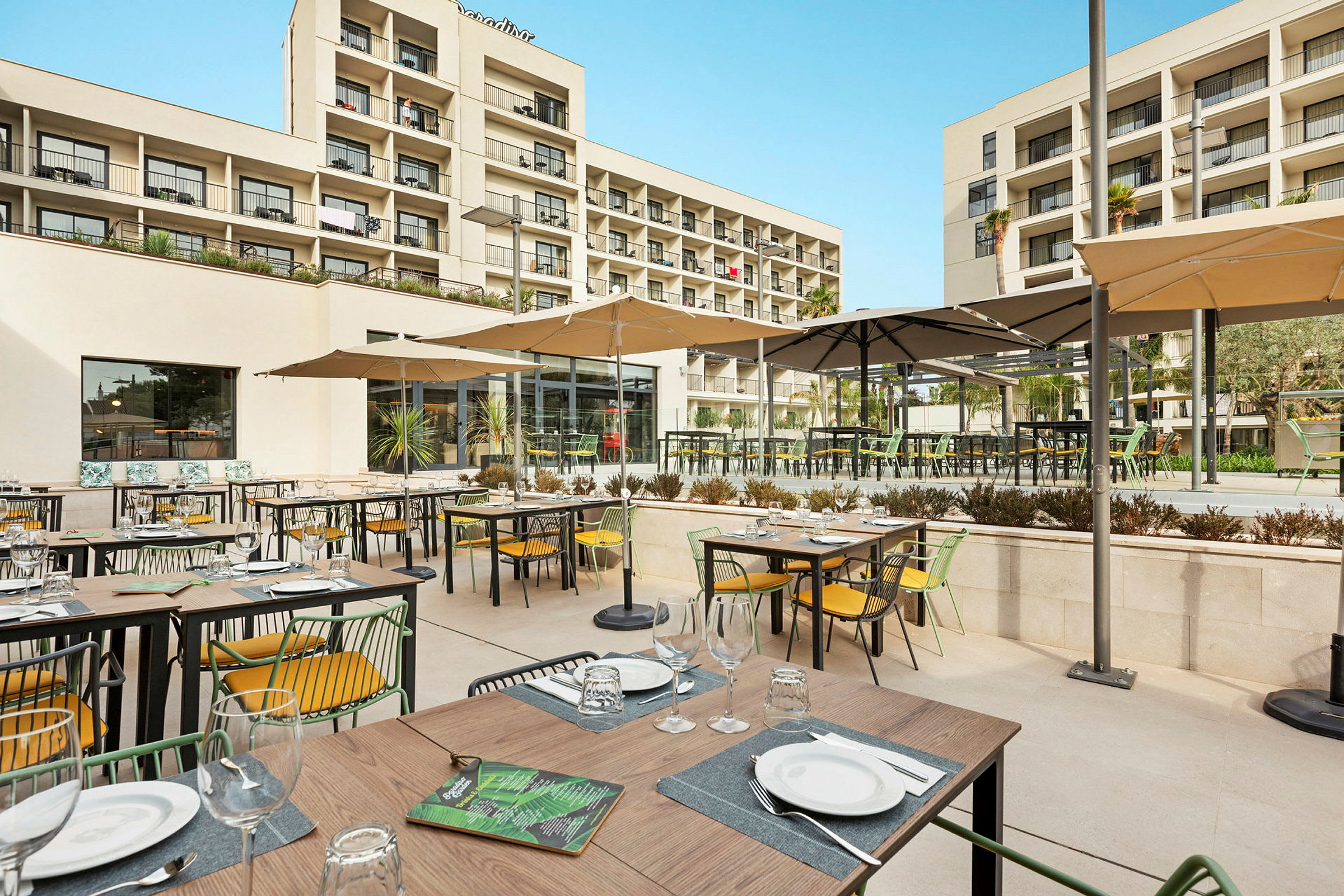 Bild av en utomhusrestaurang med uppställda bord och stolar under parasoller, samt hotellbyggnaden i bakgrunden. (AI-genererad text)