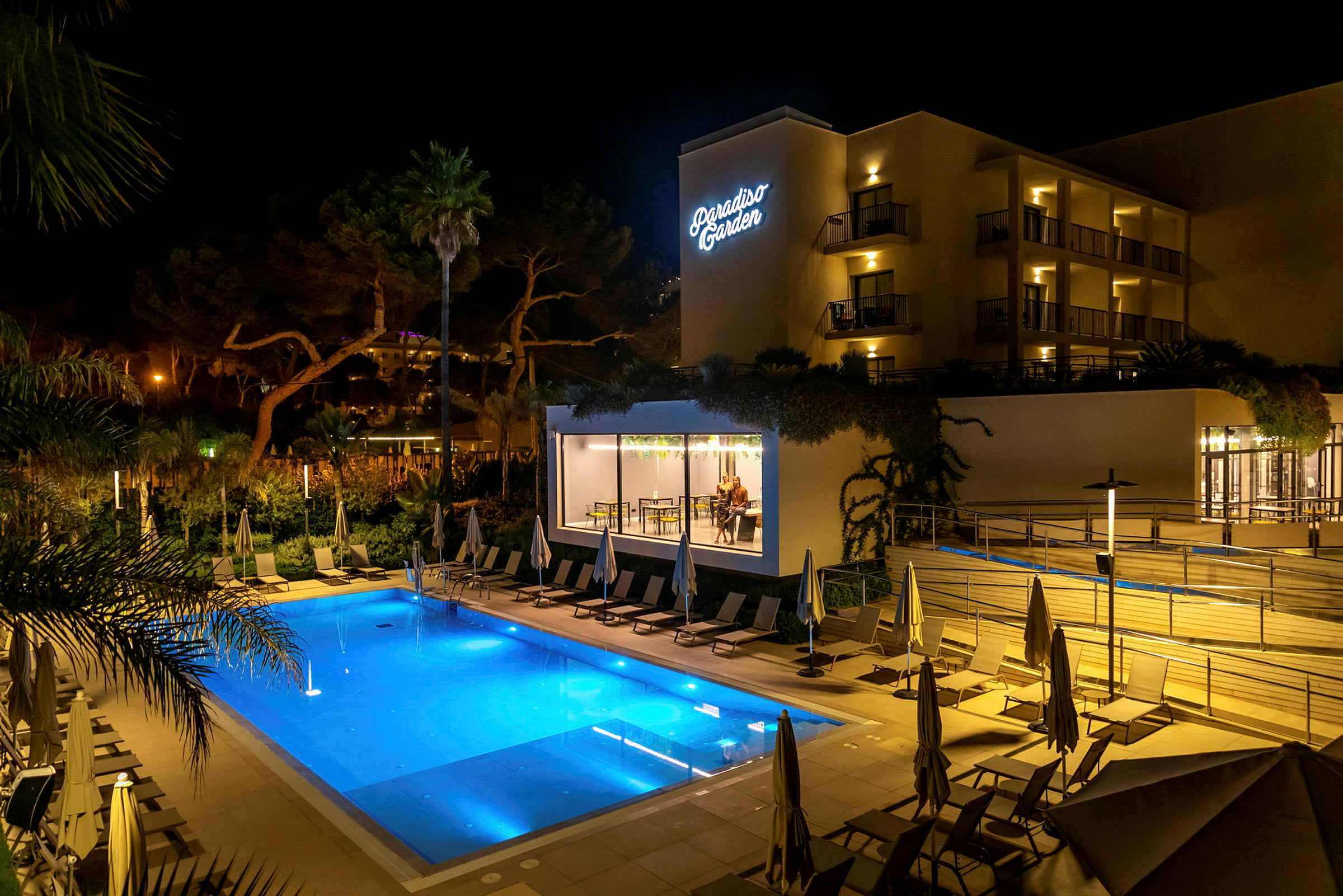 Hotell Paradiso Garden sett på kvällen med en upplyst pool och solstolar i förgrunden. (AI-genererad text)