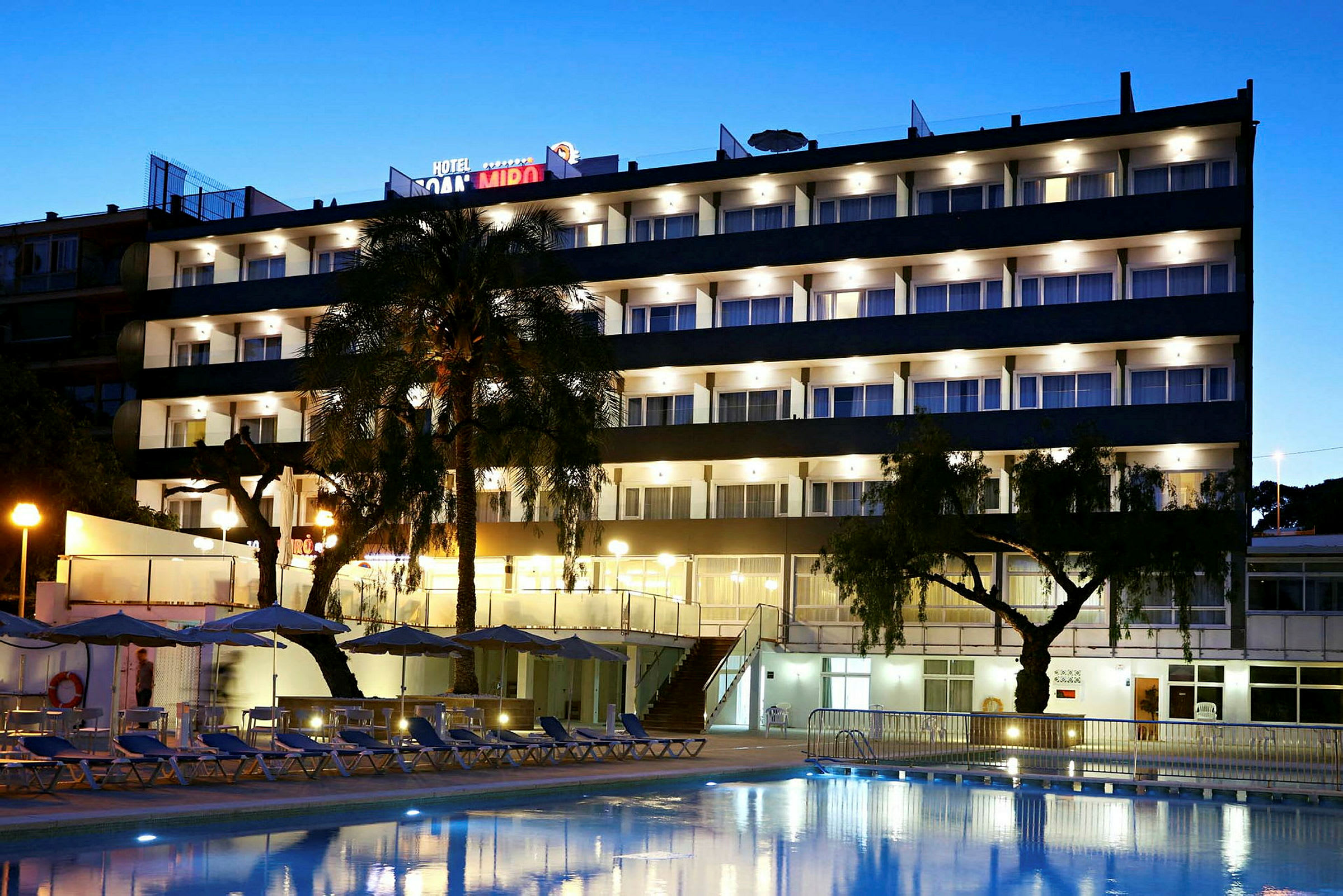 Ett hotell i flera våningar med balkonger och upplysta fönster, omgivna av träd, parasoller och en pool i förgrunden. (AI-genererad text)