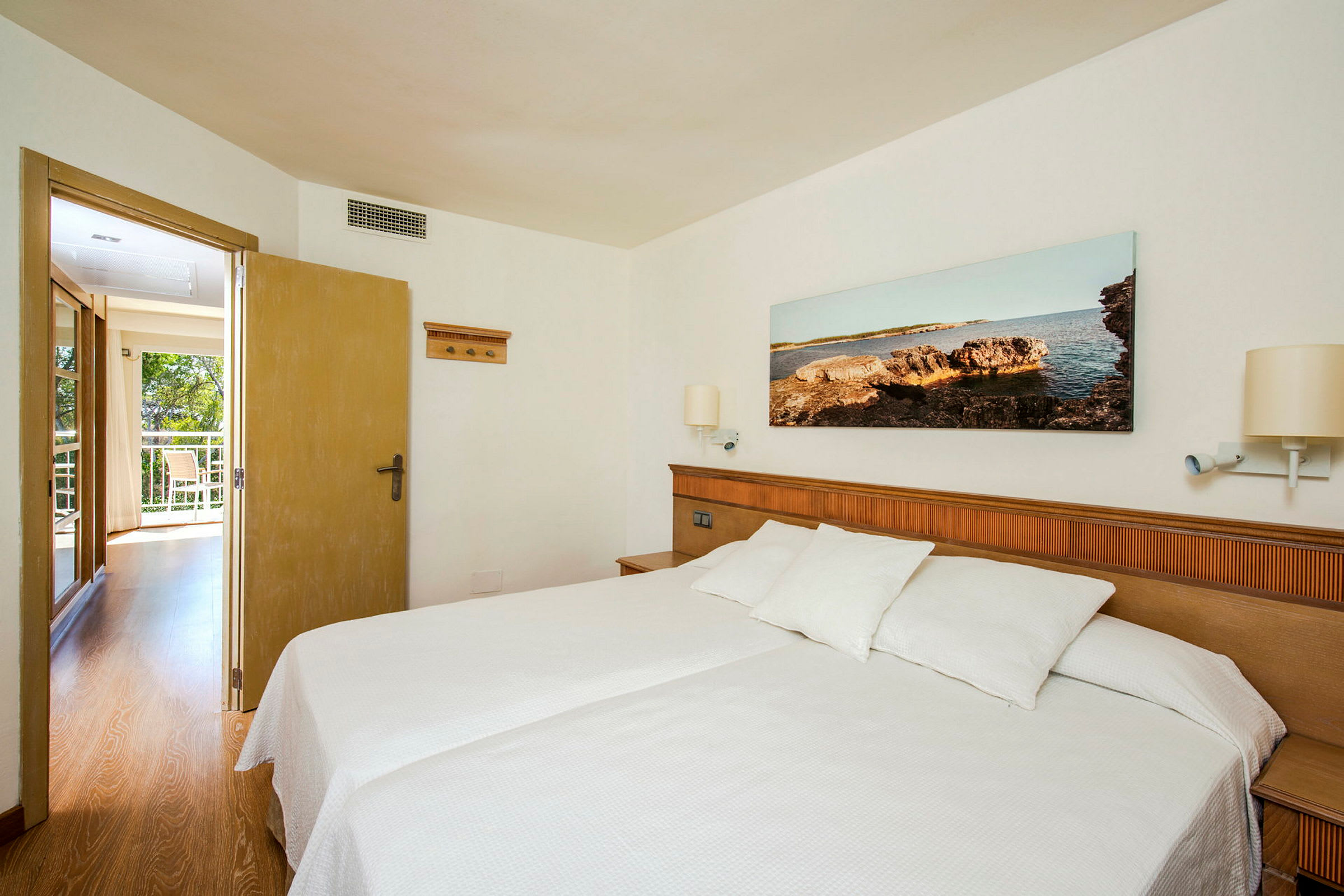 Ett hotellrum med en dubbelsäng och vita sängkläder. På väggen ovanför sängen hänger en tavla med ett kustmotiv. Dörren till höger leder till en ljus och öppen balkong. (AI-genererad text)