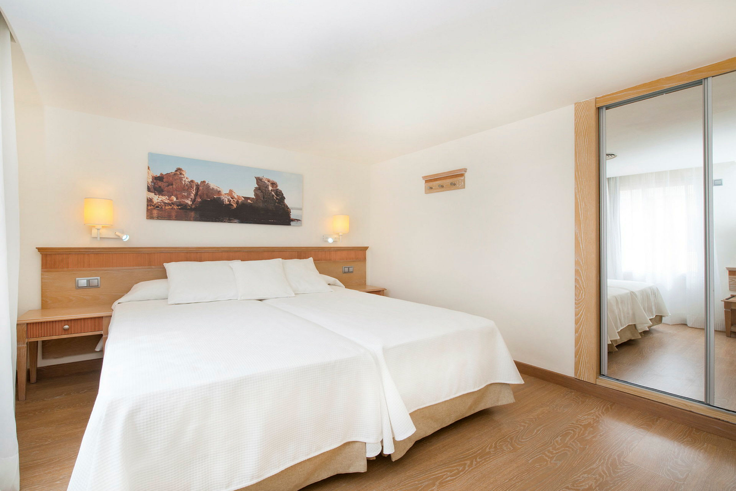 Ett hotellrum med två enkelsängar täckta med vita sängkläder, en träpanel bakom sängarna med bordslampor, spegeldörrar på en garderob och ett stort fönster som släpper in naturligt ljus. (AI-genererad text)