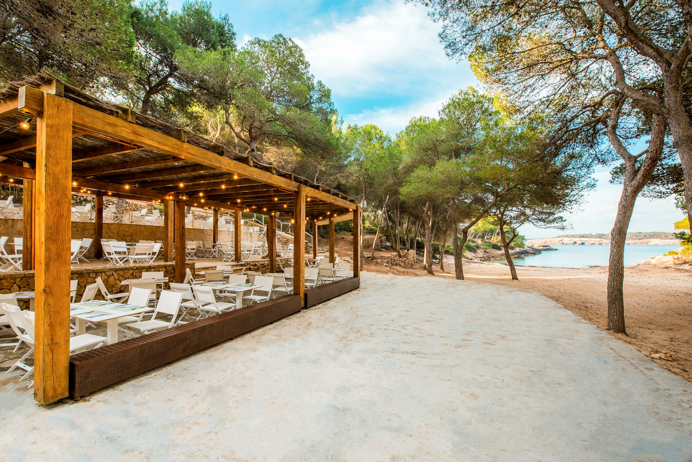 En strandrestaurang med vita möbler och en träpergola nära sandstrand och träd. (AI-genererad text)