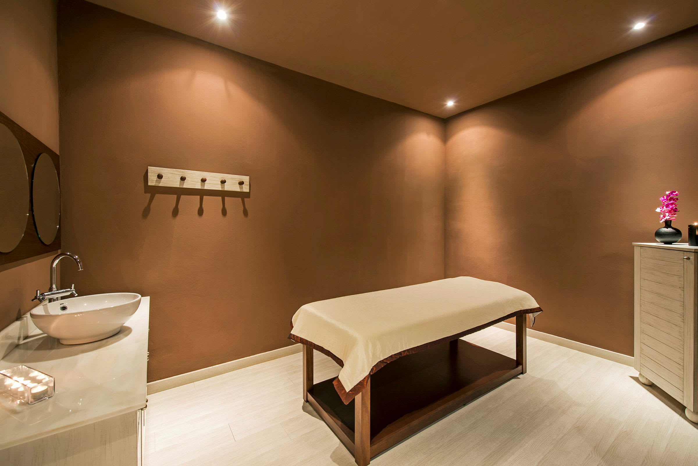 Ett spa-rum med en massagebänk, en diskbänk med spegel och en skänk med en dekoration. (AI-genererad text)