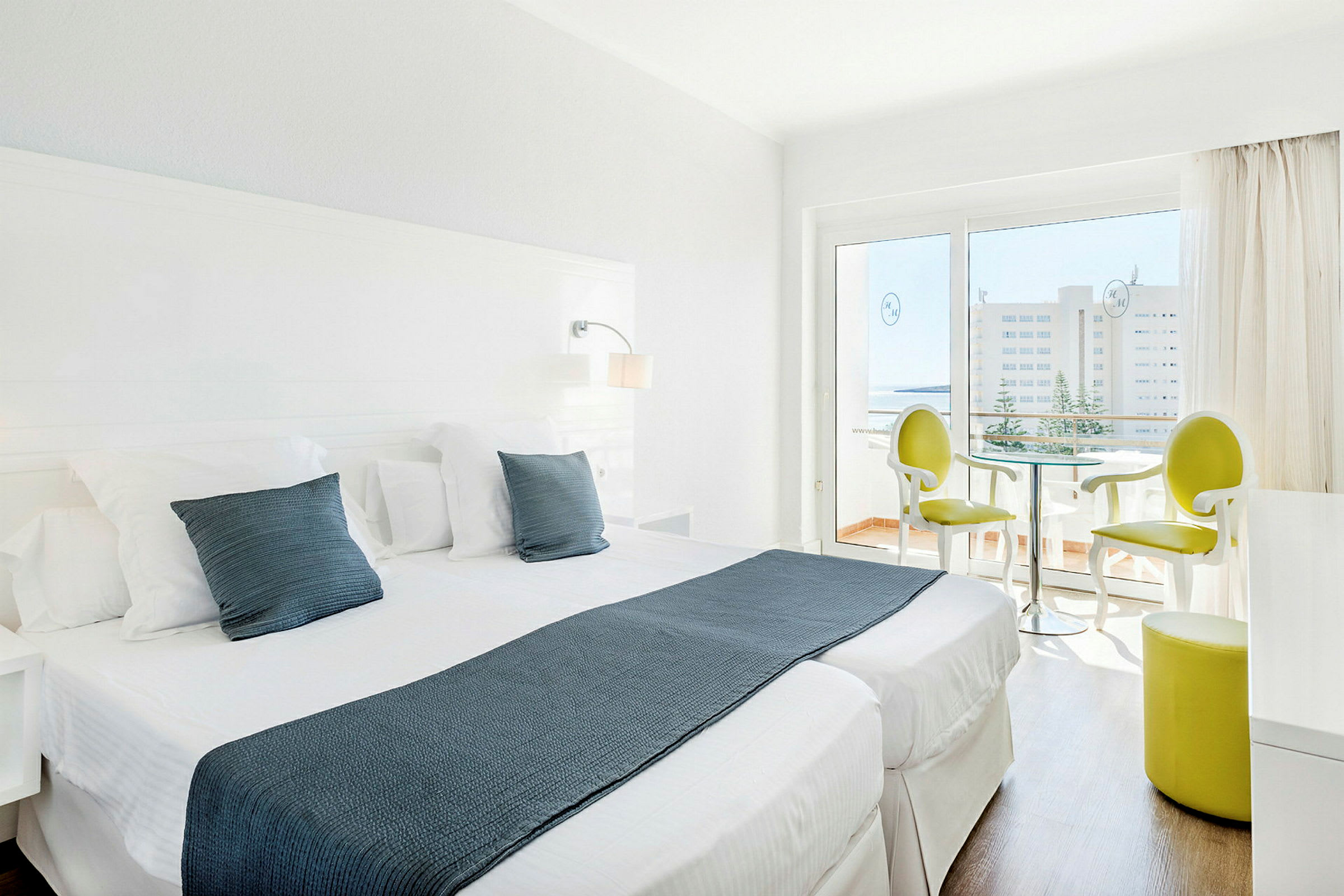 Ett ljust hotellrum med två enkelsängar, vita möbler och en sittgrupp nära en glasdörr som leder till en balkong. (AI-genererad text)