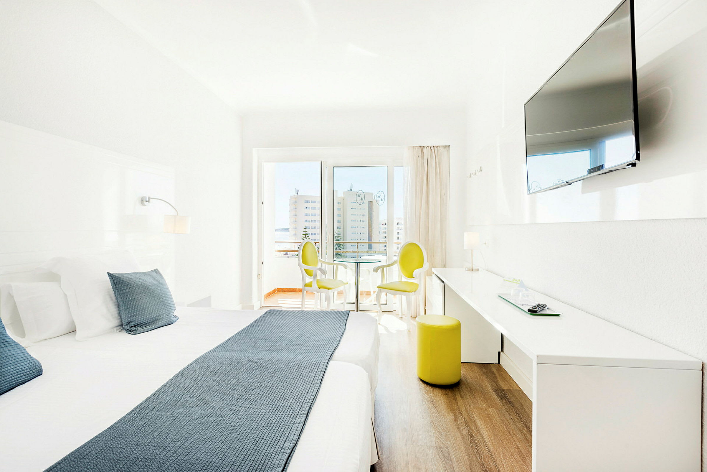 Ljust hotellrum med dubbelsäng, TV på väggen och sittmöbler vid fönster. (AI-genererad text)