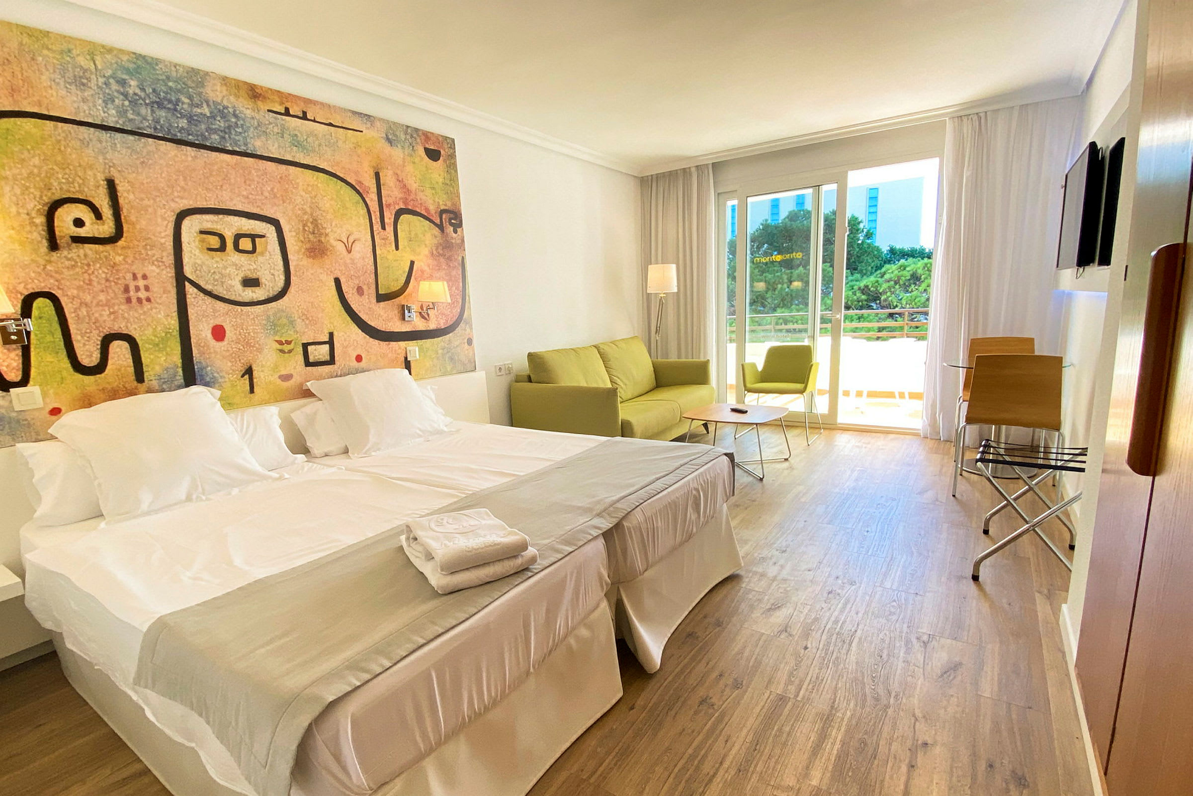 Ett hotellrum med dubbelsäng, färgglad väggkonst, och en sittgrupp med soffa och stol vid fönstret. (AI-genererad text)