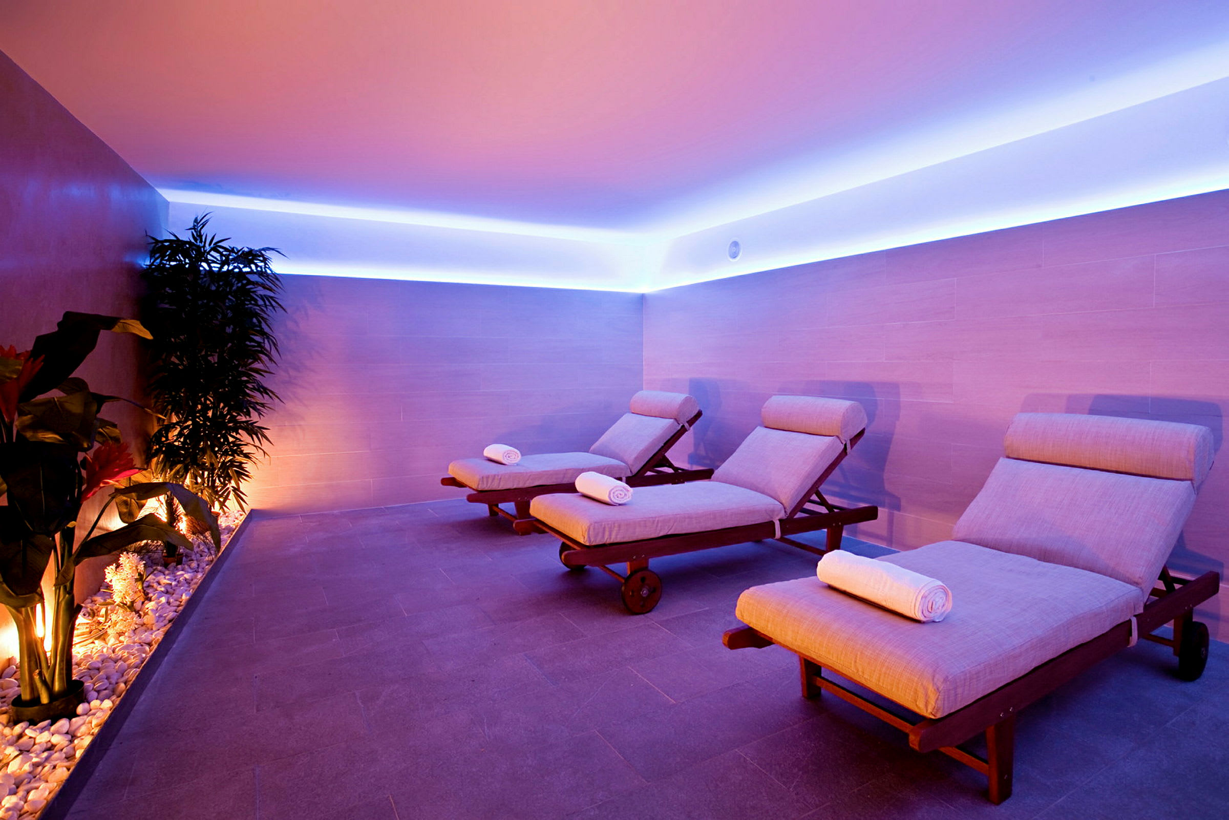 Ett spa-rum med tre solstolar med kuddar och vikta handdukar, omgivna av mjuk belysning och växter. (AI-genererad text)