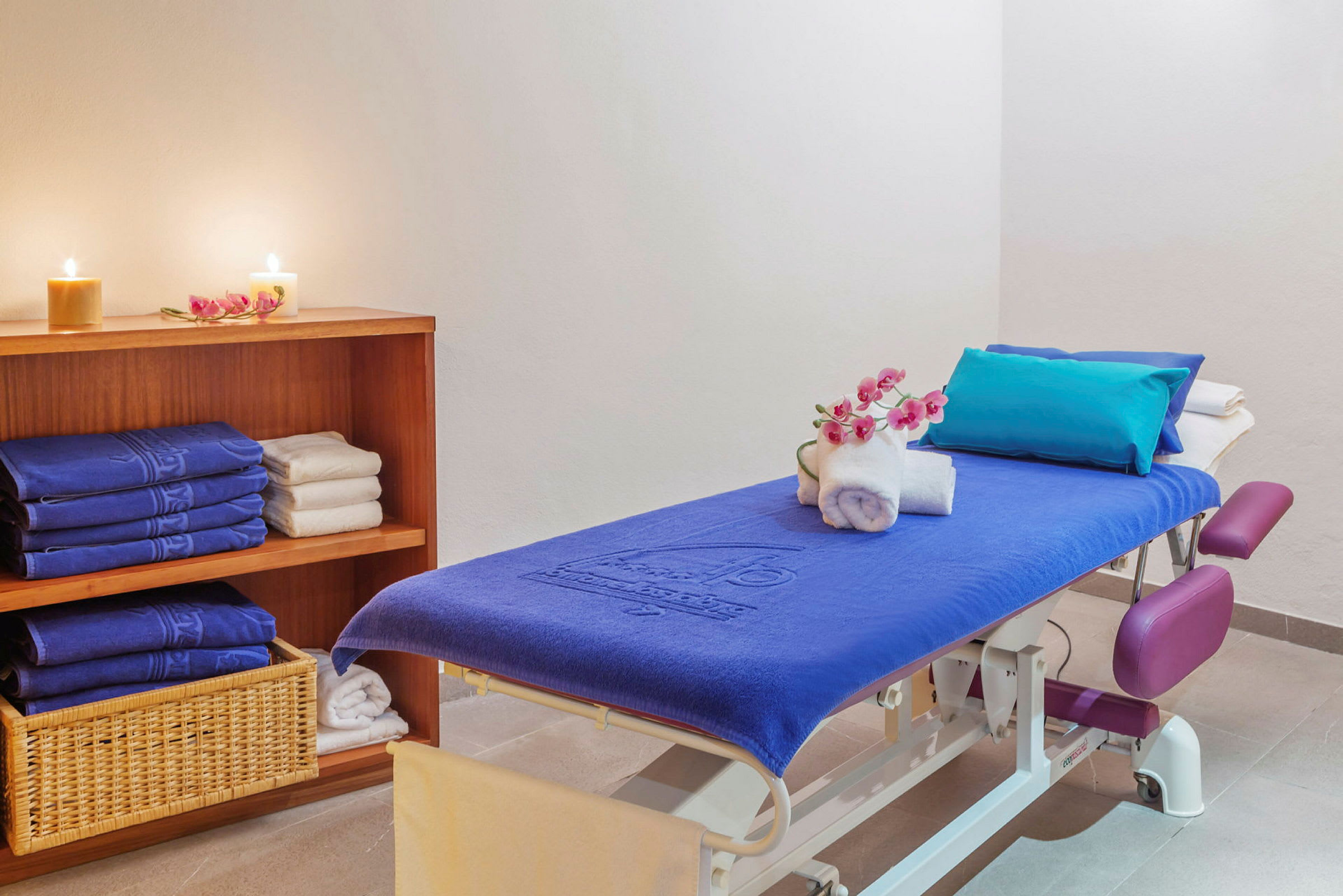 Ett massagesrum med en massagebänk klädd i en blå handduk, dekorerad med vita handdukar och en blomsterkvist. I bakgrunden finns en hylla med vikta handdukar och tända ljus. (AI-genererad text)