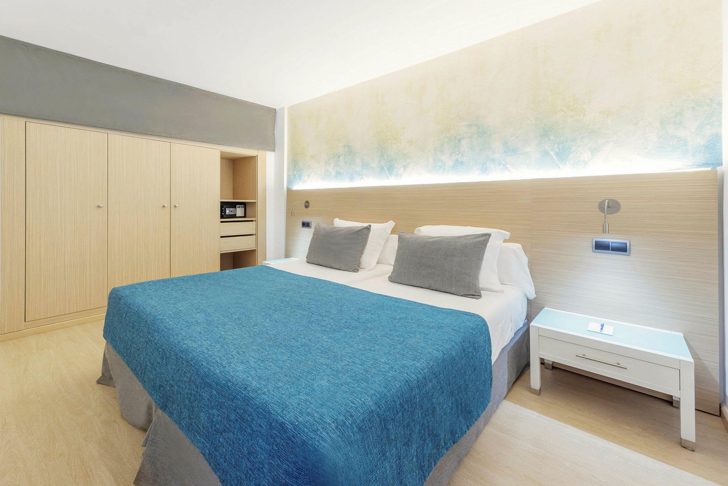 Hotellrum med dubbelsäng, blå sängöverkast, vita kuddar och minimalistisk inredning. (AI-genererad text)