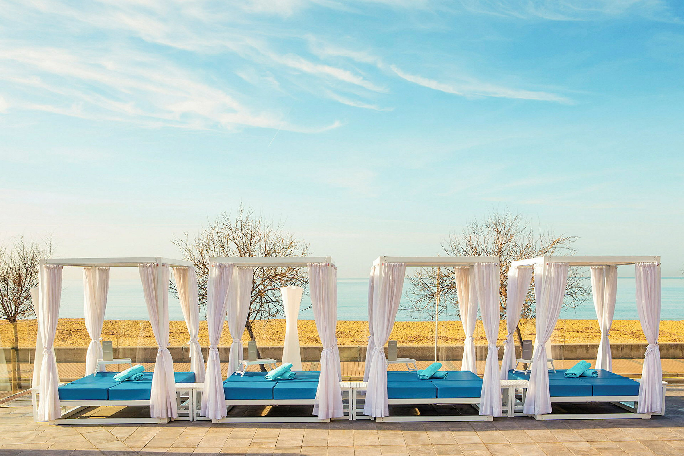 Liggande cabanas med vita draperier och blå dynor framför stranden. (AI-genererad text)