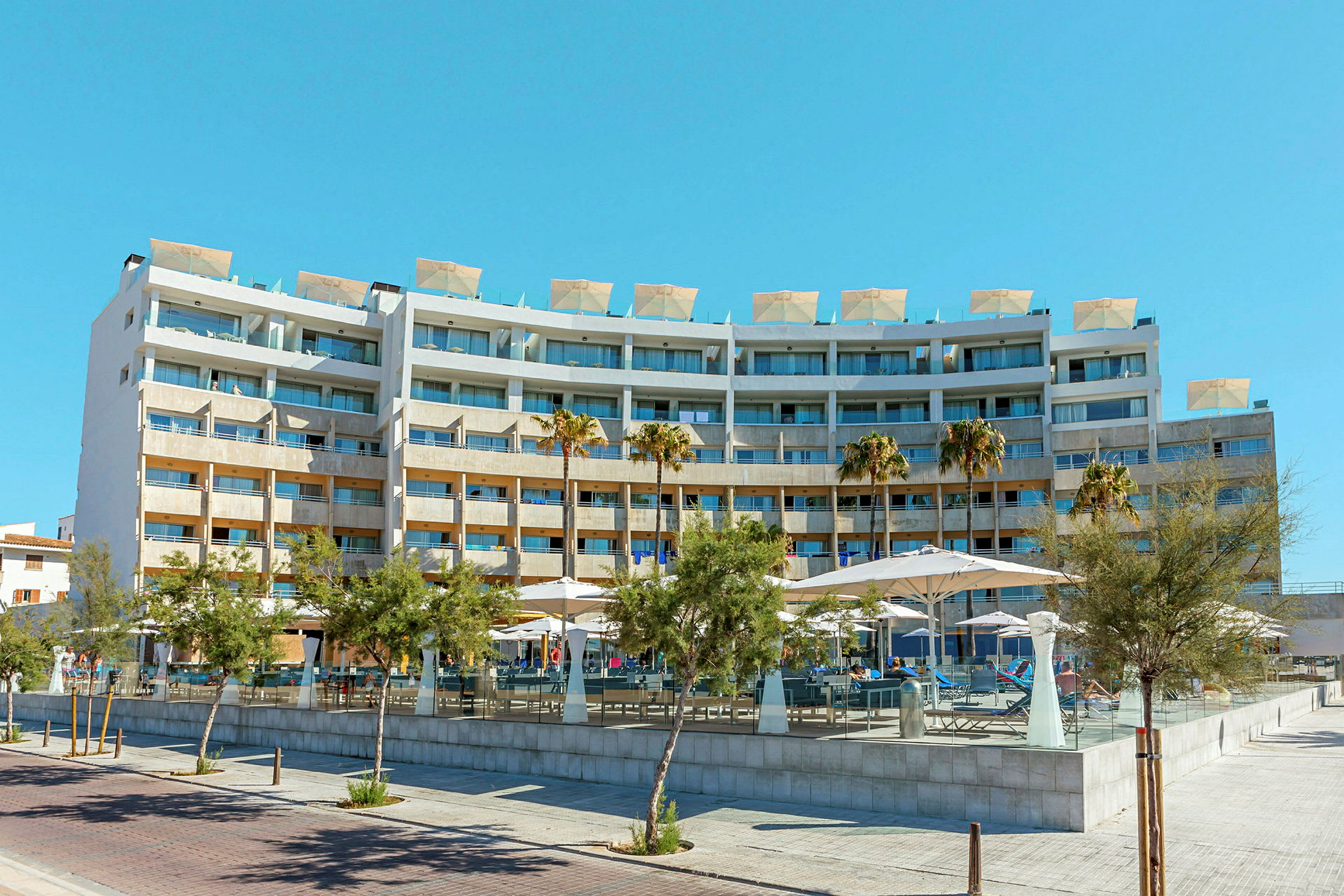 Ett större hotellkomplex med flera våningar och balkonger. Framför byggnaden finns några träd och parasoller. (AI-genererad text)