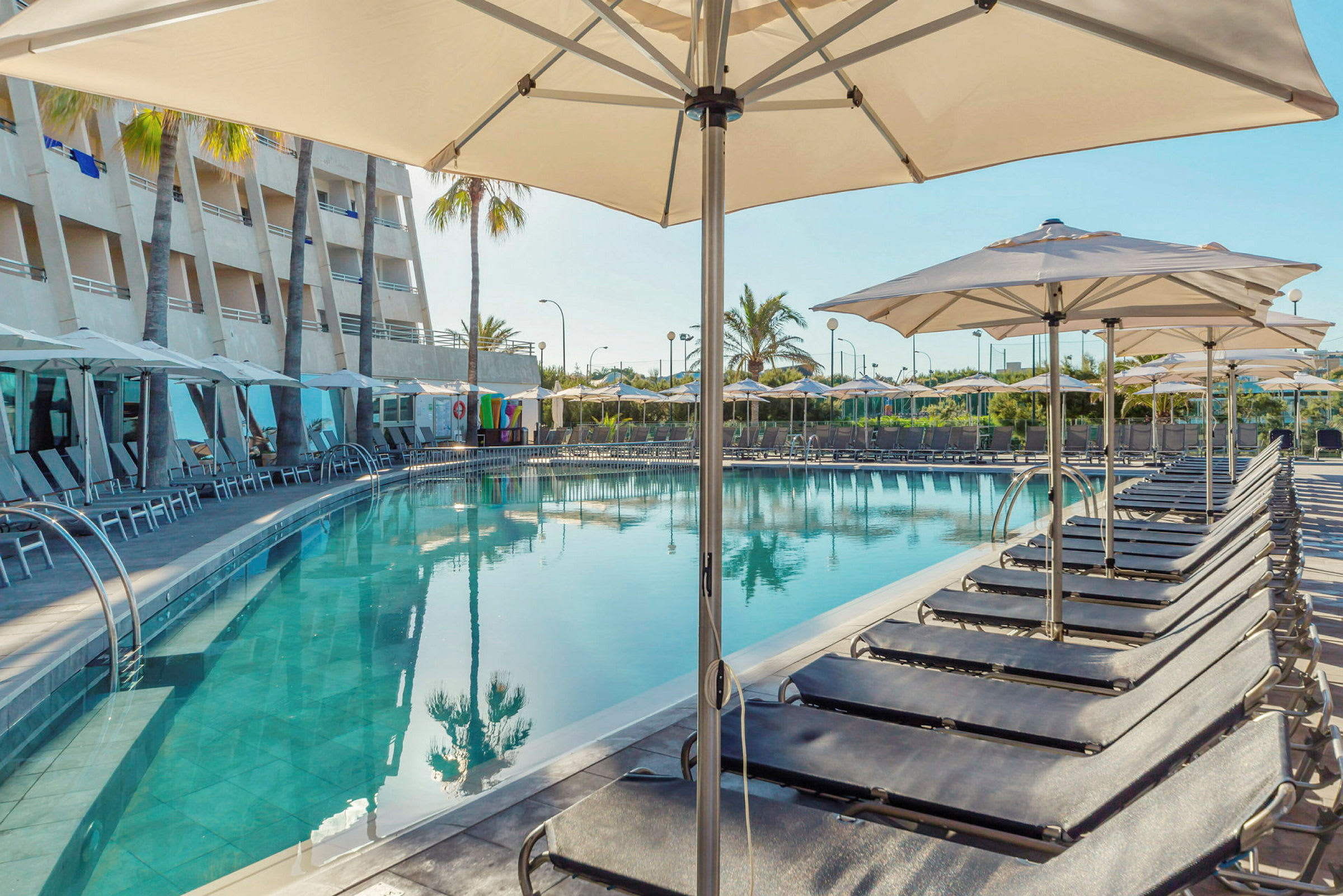 Hotellpool med solstolar och parasoller vid sidan, samt hotellbyggnad och palmer i bakgrunden. (AI-genererad text)