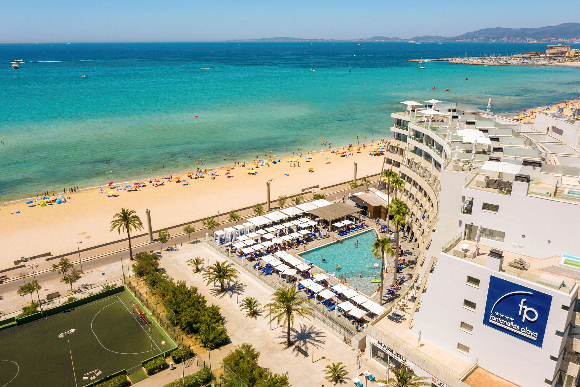 Vy över en strand med klart turkosblått hav och en hotellresort med pool i förgrunden. (AI-genererad text)