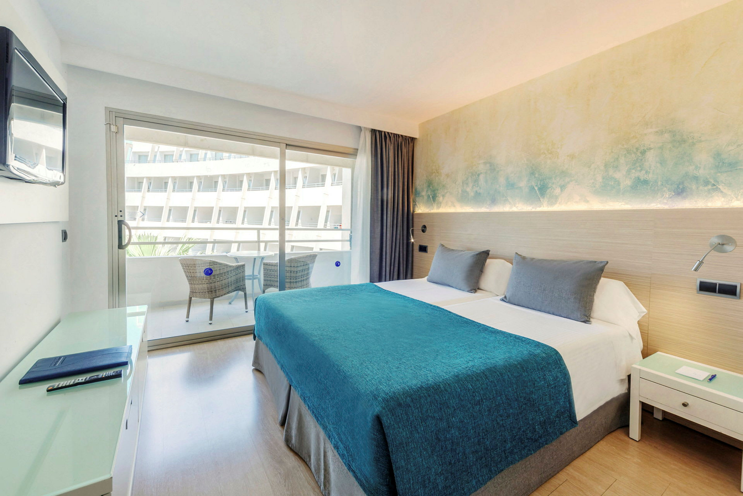 Ett hotellrum med en dubbelsäng, blått överkast och nattduksbord. En balkong med sittmöbler ses genom glasdörren. (AI-genererad text)