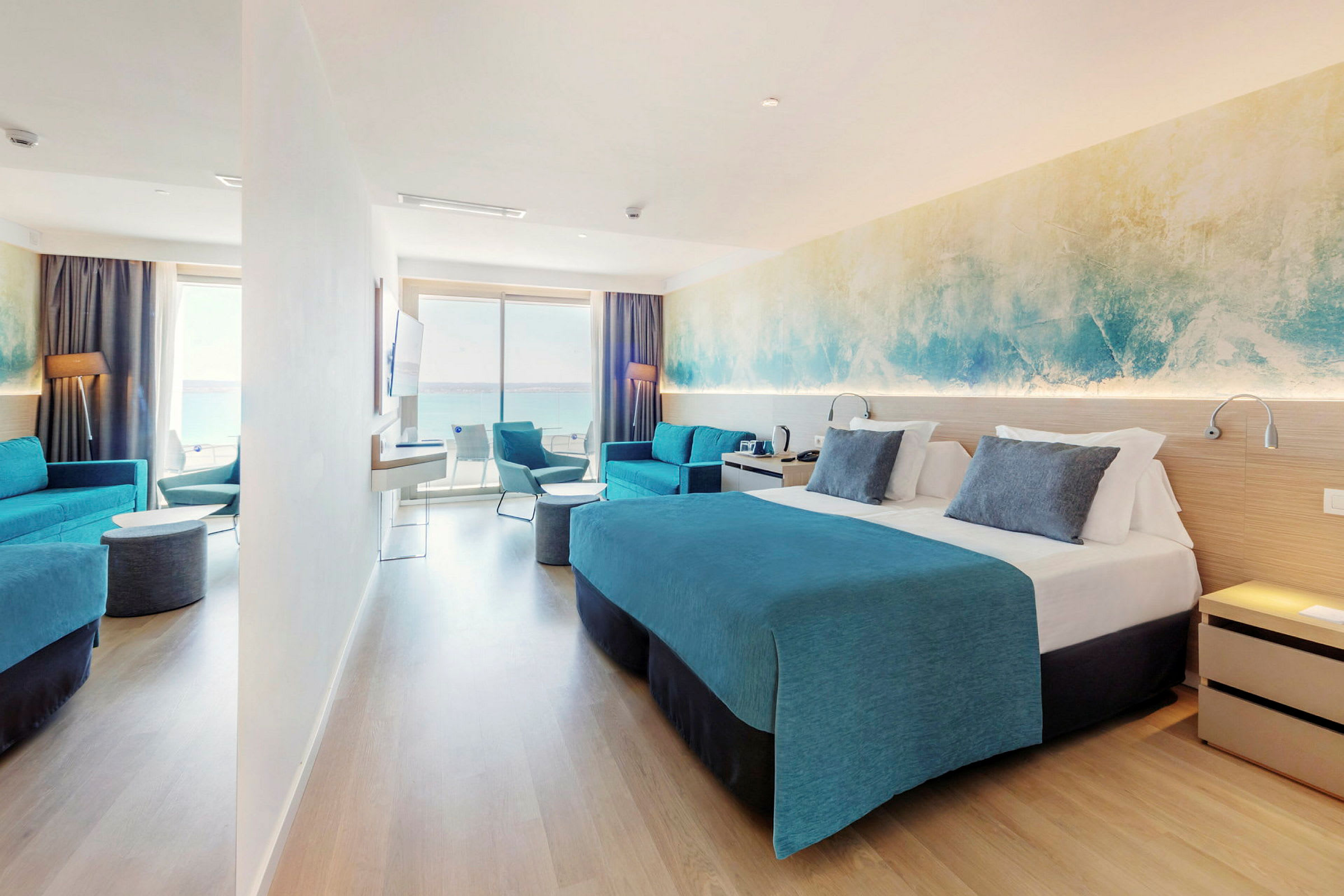 Ett hotellrum med dubbelsäng, blå inredning och en sittgrupp. Ett stort fönster ger utsikt över havet. (AI-genererad text)