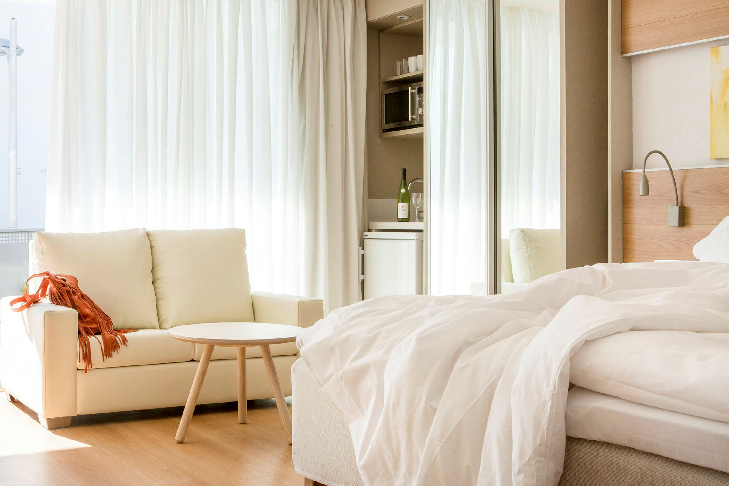 Ett ljust hotellrum med en soffa, ett litet bord och en bäddad säng. I bakgrunden finns ett minikök med kylskåp. (AI-genererad text)