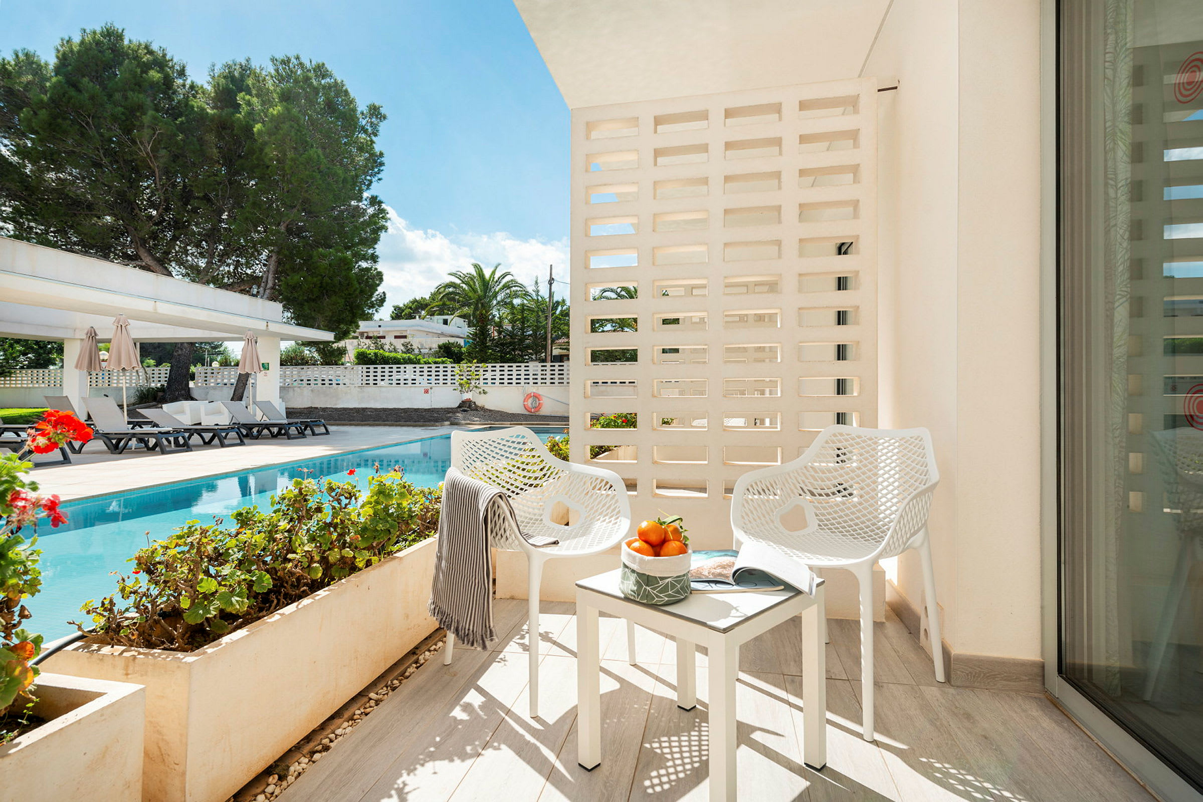 Bild av en balkong med två vita stolar och bord med apelsiner, med utsikt över en pool och trädgård. (AI-genererad text)
