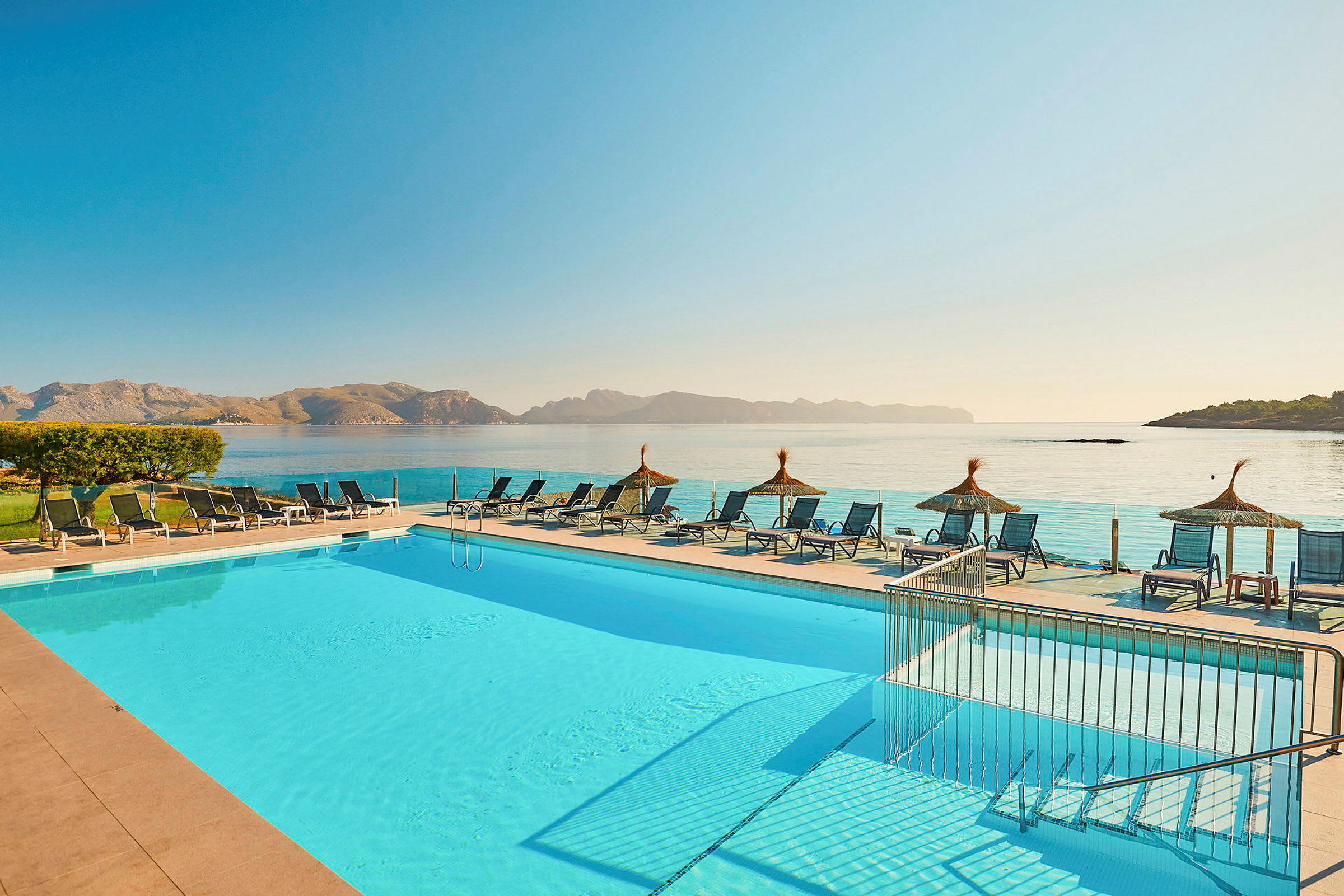 En pool med solstolar och utsikt över havet samt berg i bakgrunden. (AI-genererad text)