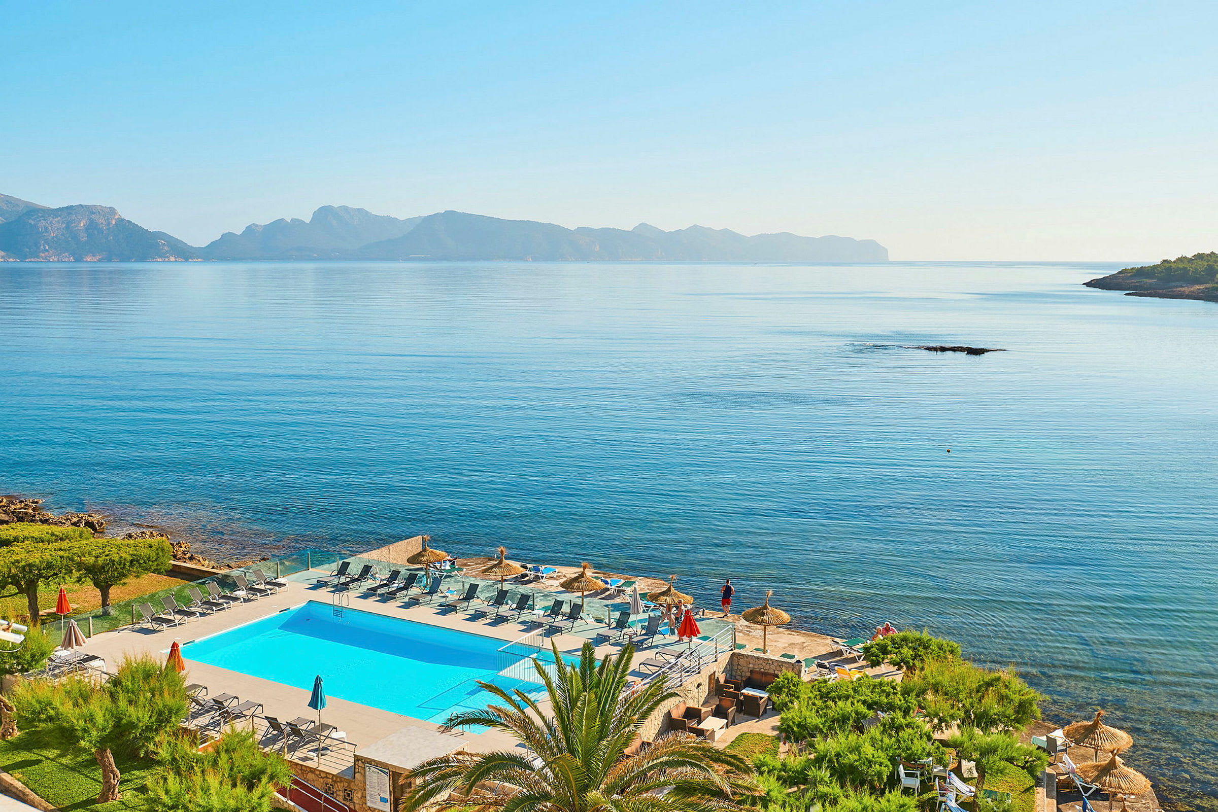 Utsikt över en pool med solstolar och parasoller vid havet samt berg i bakgrunden. (AI-genererad text)