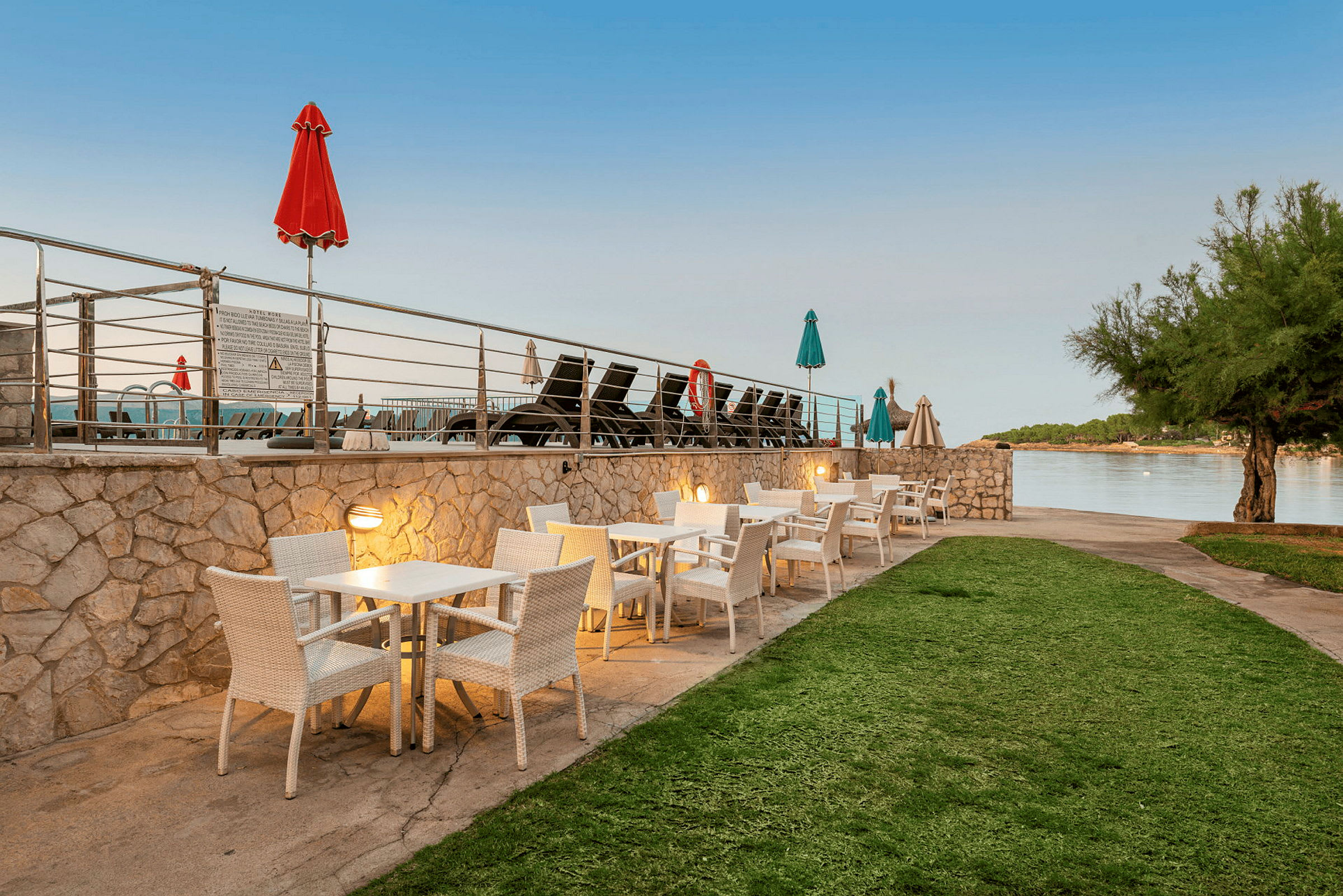 Utomhusbord med vita stolar vid en stenmur, nära en pool och havet. (AI-genererad text)
