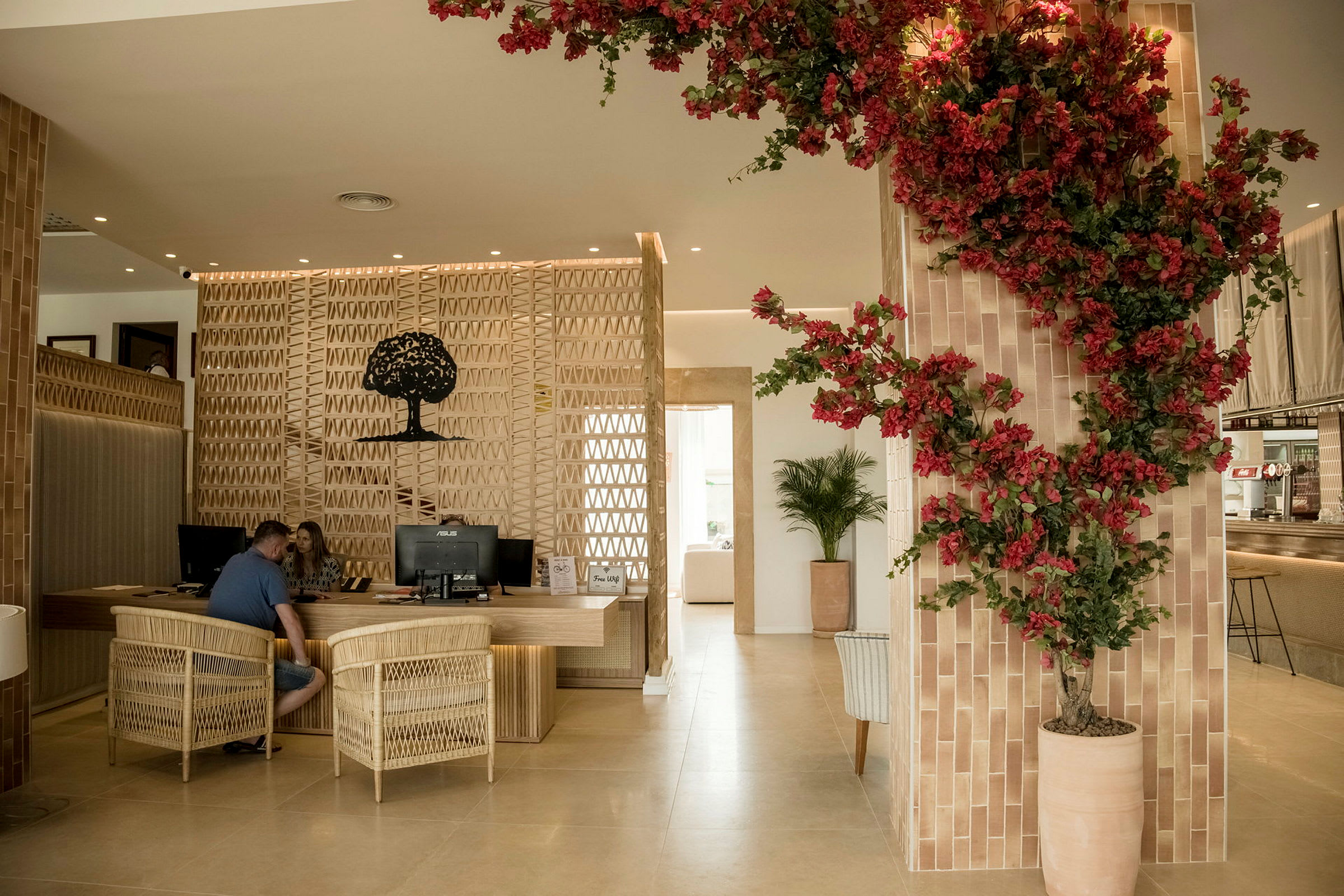 En receptionsmiljö med trämöbler och en väggdekoration av en siluett av ett träd. Röda blommor pryder en växt i en beige kruka till höger. (AI-genererad text)