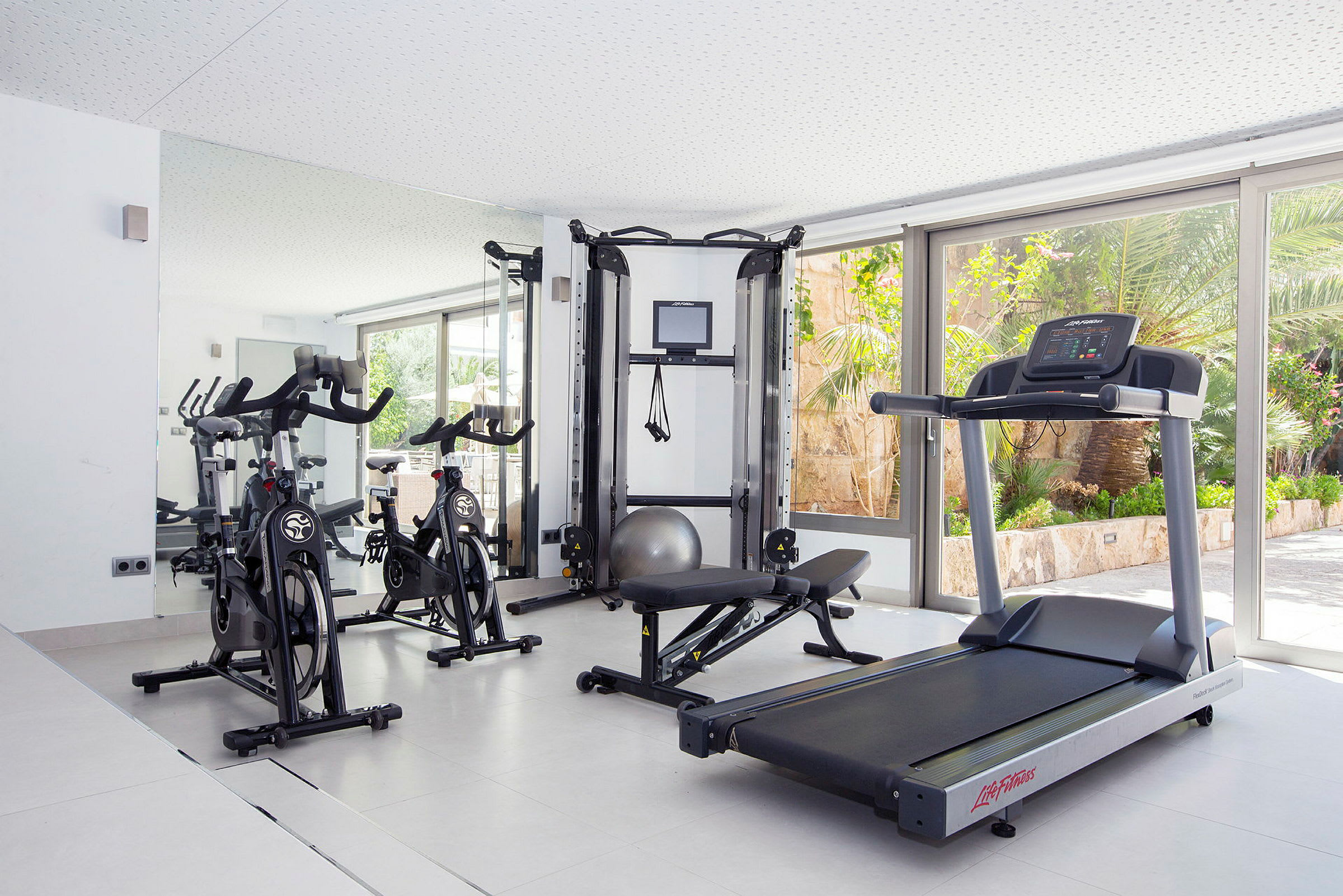 Bild av ett gym med träningsutrustning som motionscyklar, löpband och styrketräningsmaskin i ett ljust rum med stora fönster. (AI-genererad text)