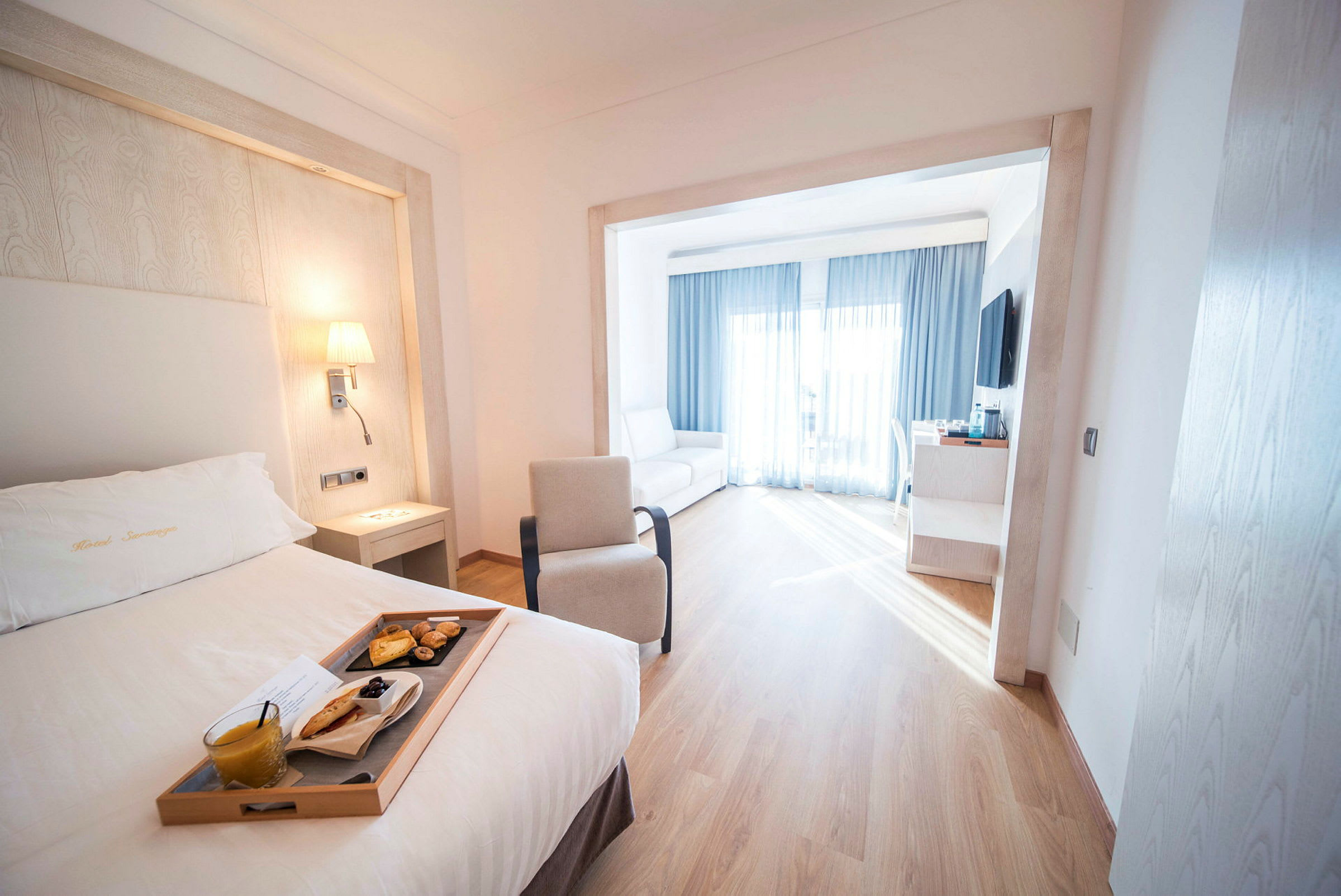Ett hotellrum med en säng och en bricka som innehåller en frukost. Rummet har ett stort fönster med ljusa gardiner. (AI-genererad text)