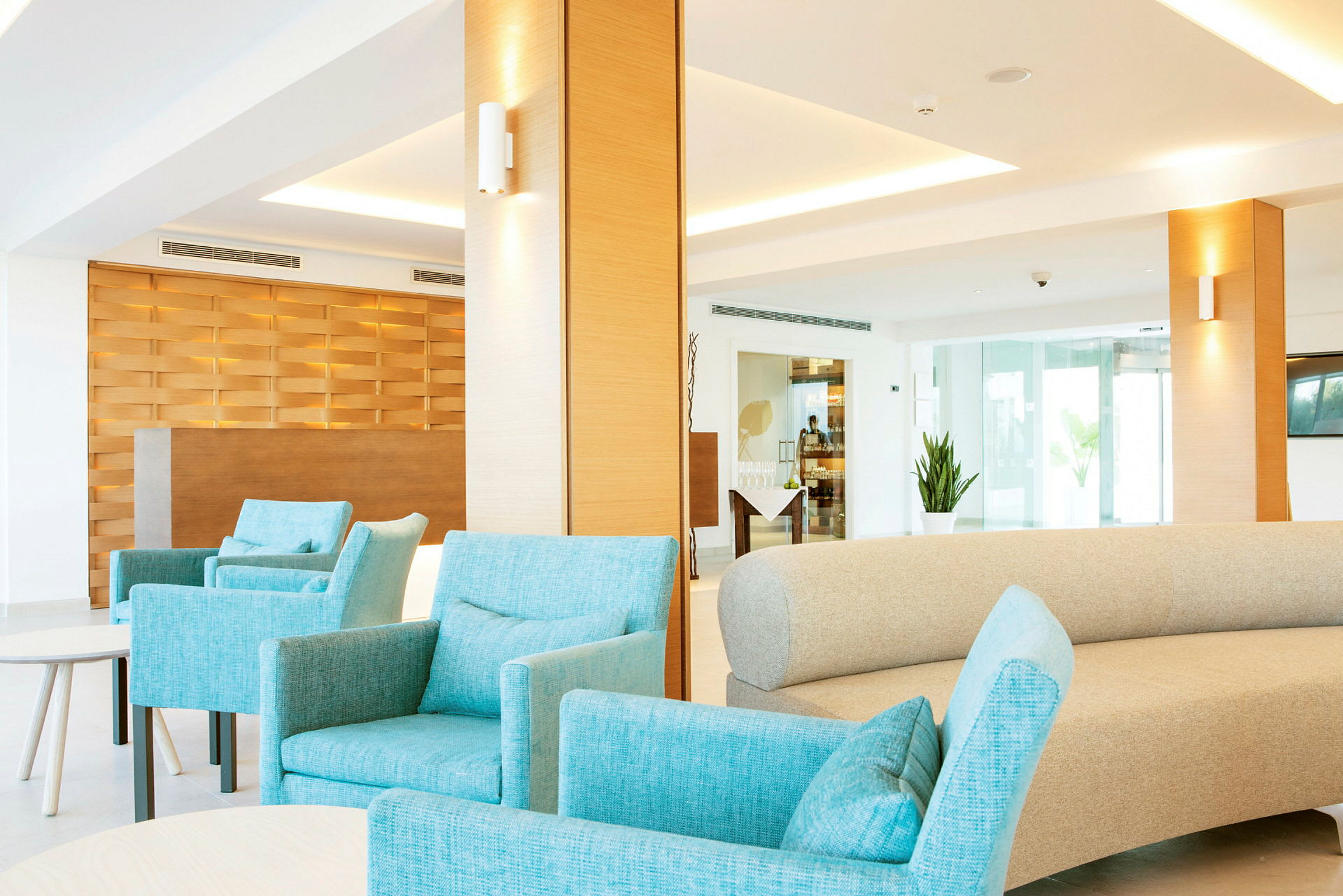 Interiör av en modern loungemiljö med turkosa fåtöljer och en beige soffa. Bakgrunden visar en reception med en träpanel och stora fönster. (AI-genererad text)
