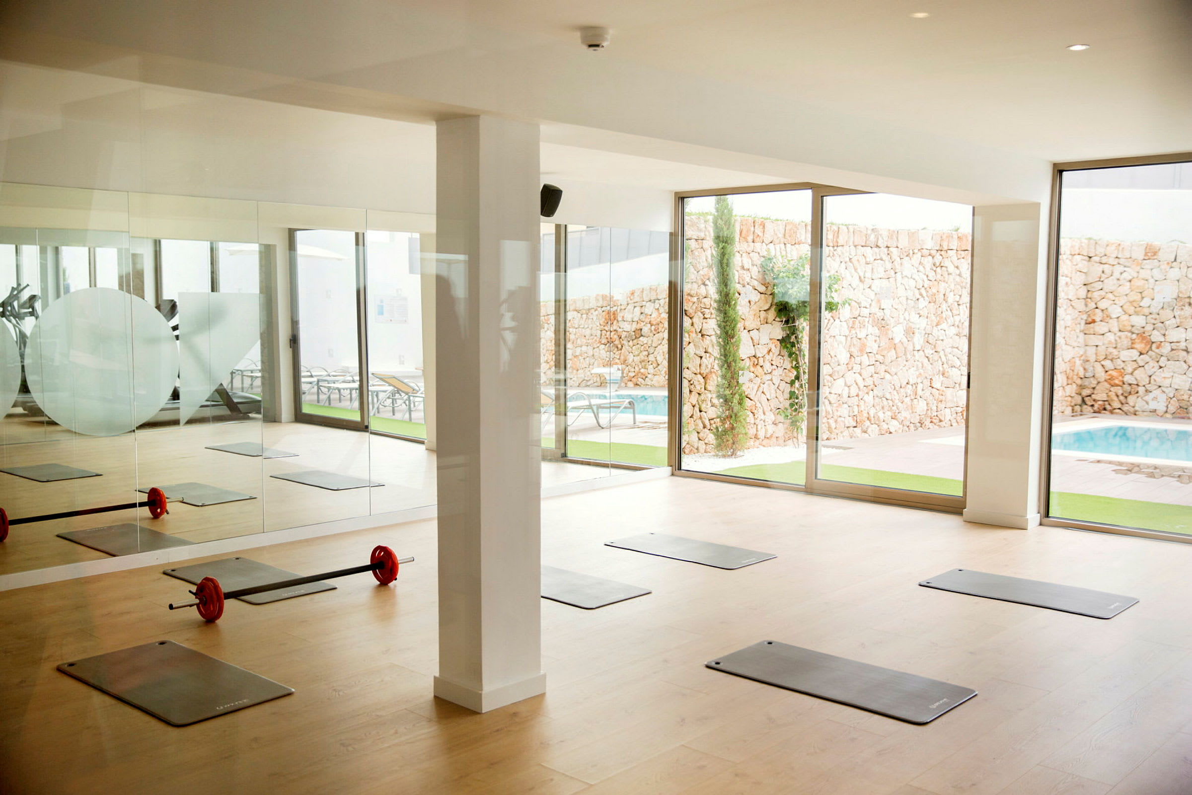 En träningssal med yogamattor och en stång med vikter på golvet nära stora glasfönster som vetter mot en pool och en stenmur. (AI-genererad text)