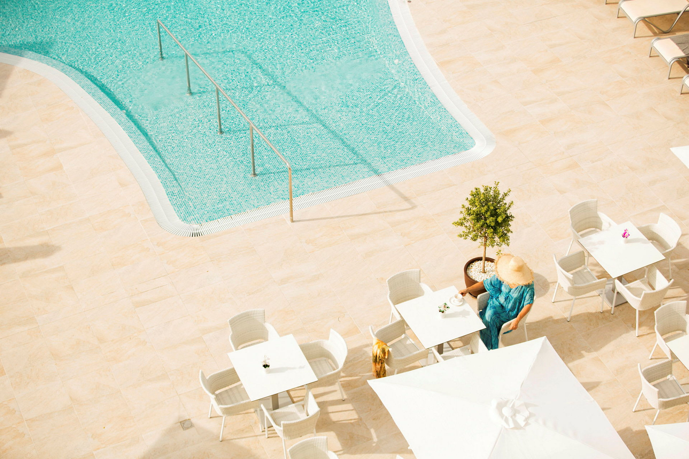 Vy över en pool med klart vatten. På en solterrass finns bord och stolar, där en person sitter vid ett bord med en kopp. Personens ansikte täcks av en solhatt. (AI-genererad text)